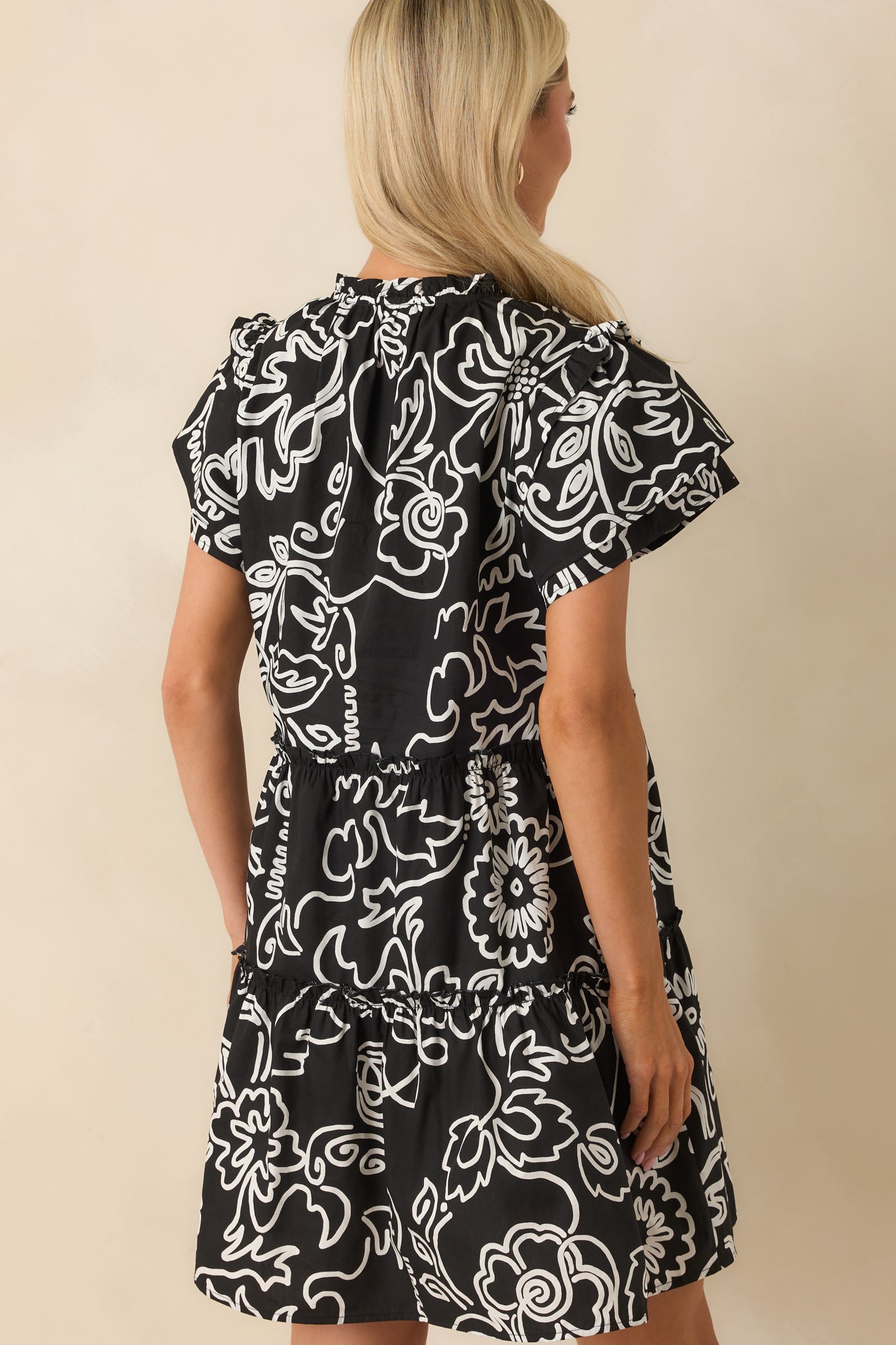 A back view of the black mini dress featuring the continued floral print, ruffle accents, and flutter sleeves from behind.