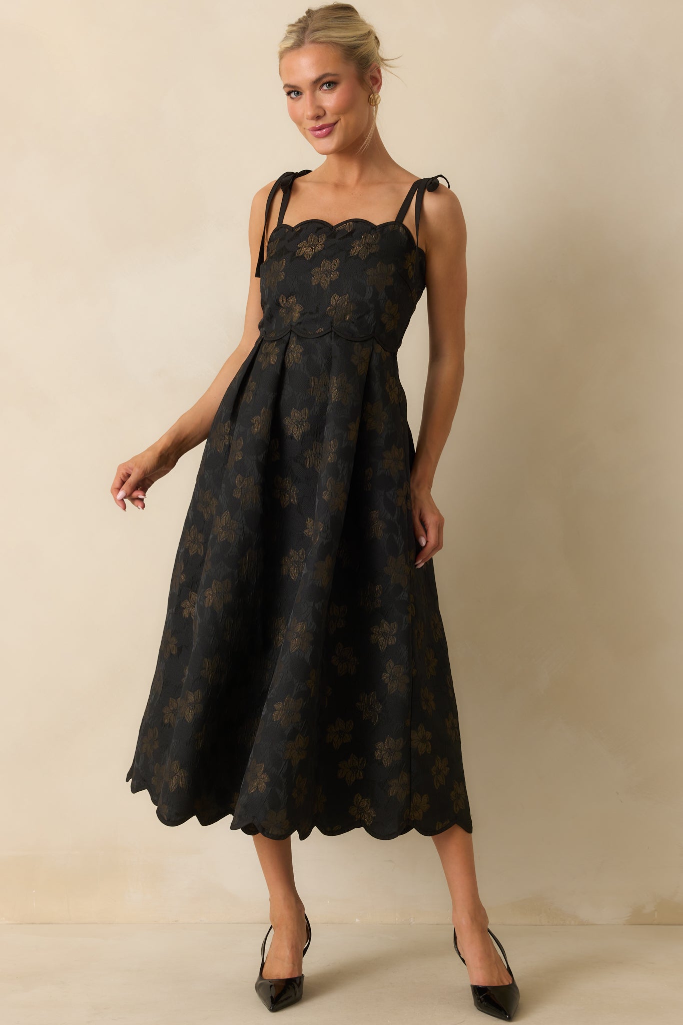 Front-facing view of black midi dress with gold floral pattern, smocked panel at the back, and tied shoulder straps, pockets visible at the sides.