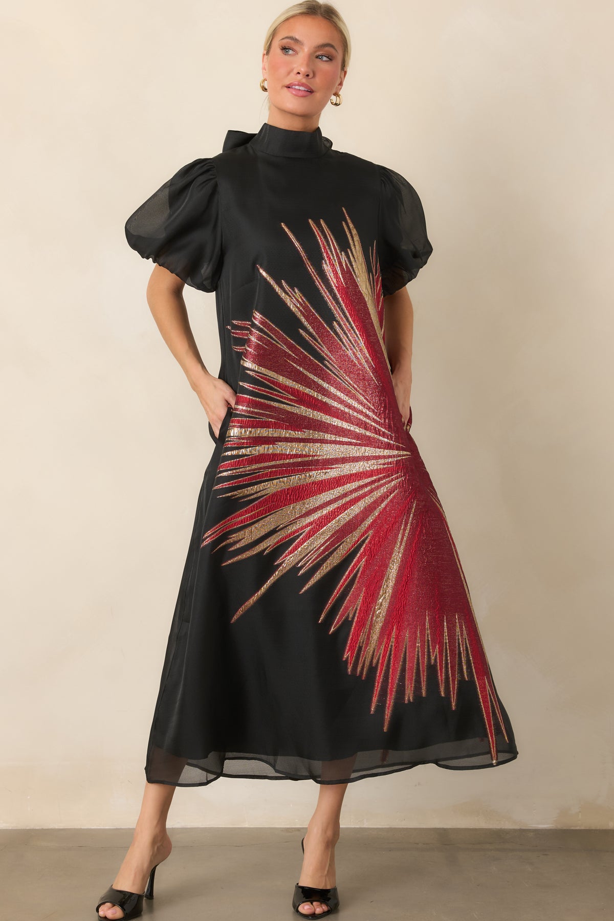 Black midi dress detailed with a dramatic metallic starburst motif, sheer sleeves, and a polished flowing skirt.