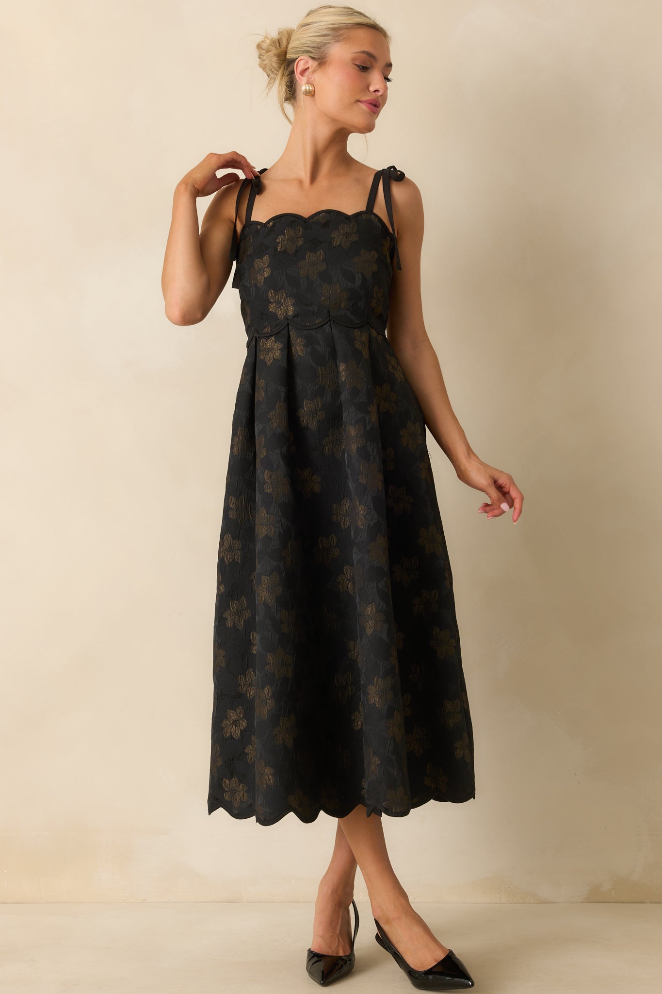 Black midi dress with scalloped hemline, self-tie shoulder straps, and subtle gold floral details, emphasizing the functional front pockets.