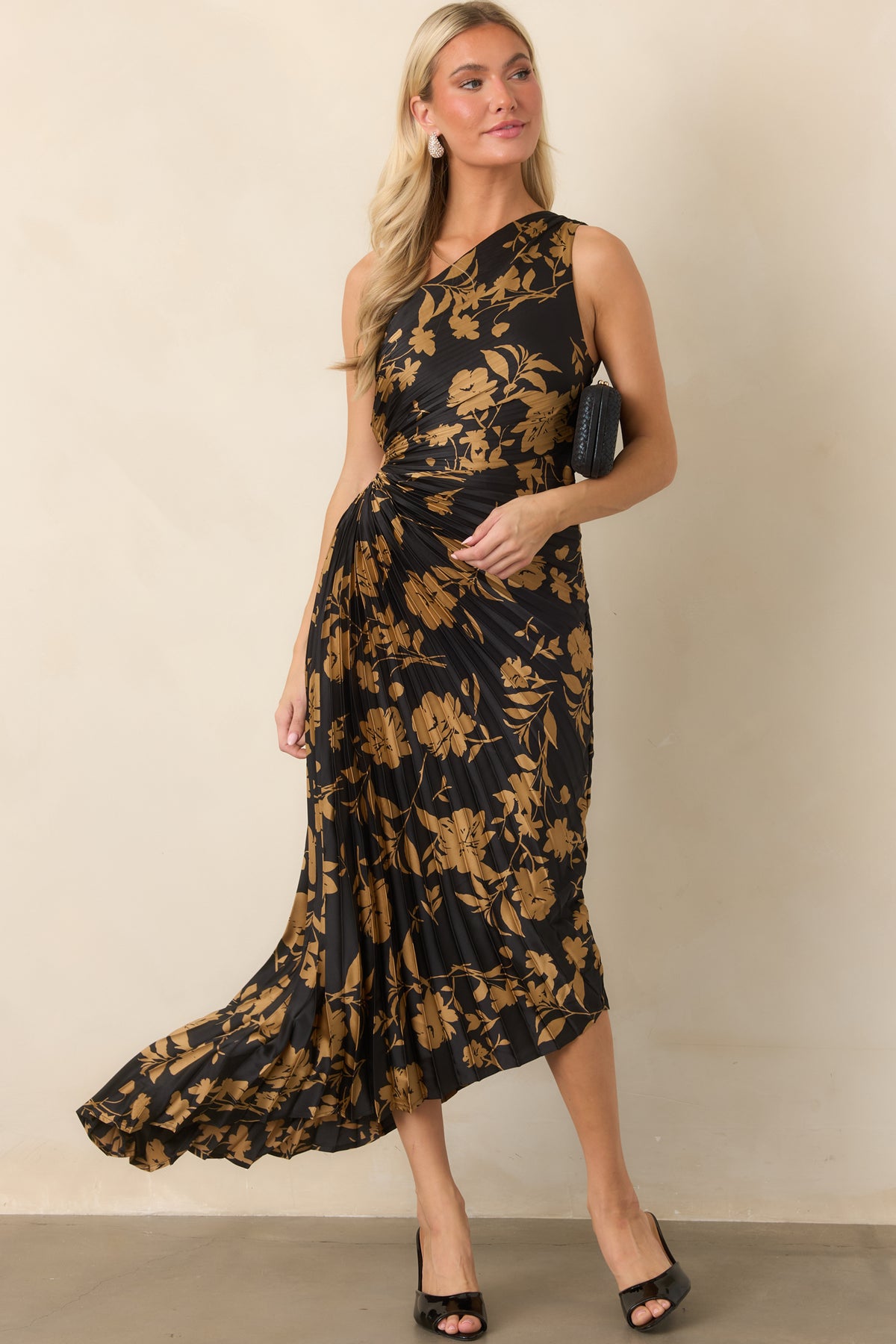 Golden floral motifs sweep across the black satin dress, highlighting its sculpted one-shoulder neckline and pleated construction.