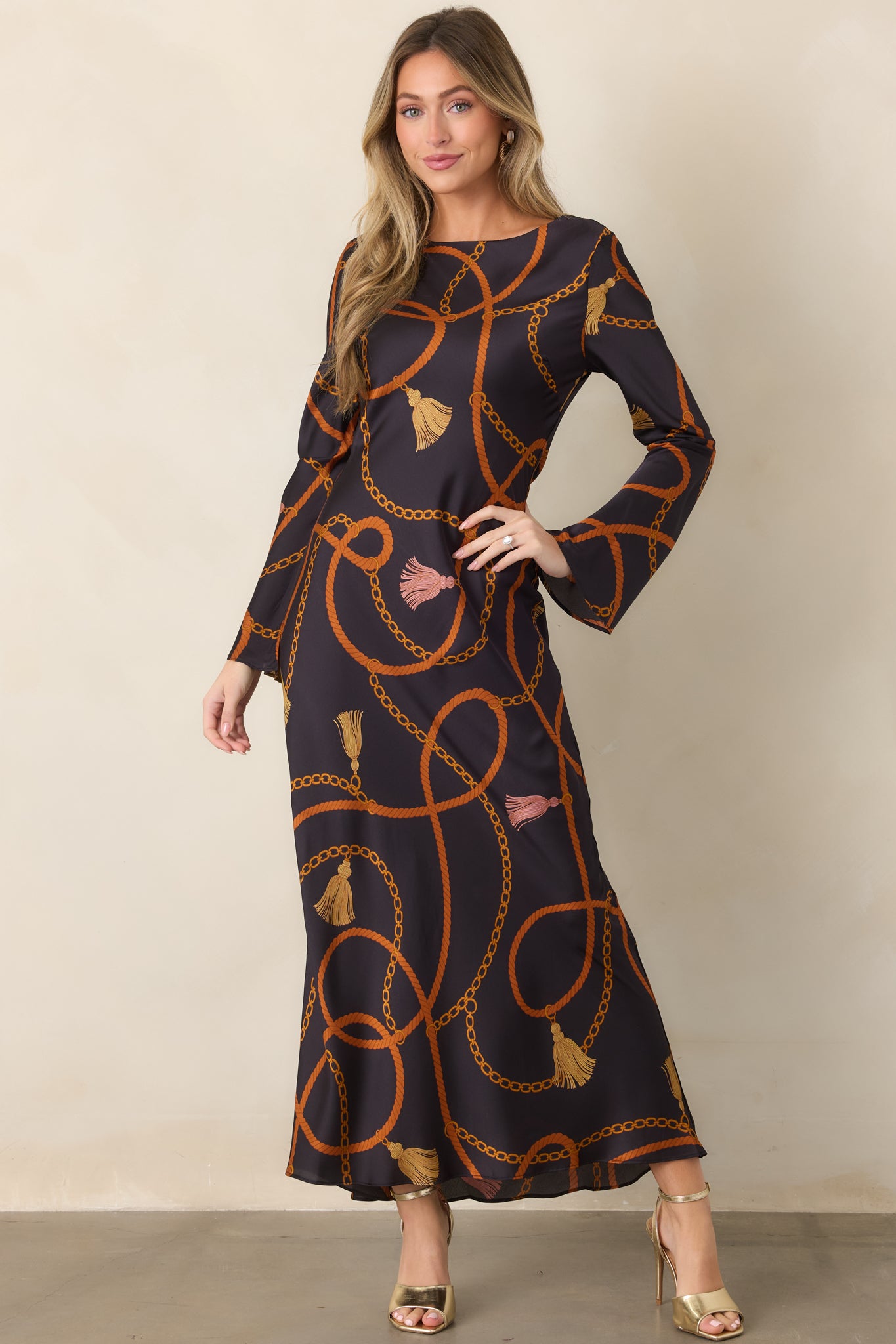 Black satin maxi dress featuring looping rust ropes, golden chains, and tassel accents with a round neckline and long sleeves.