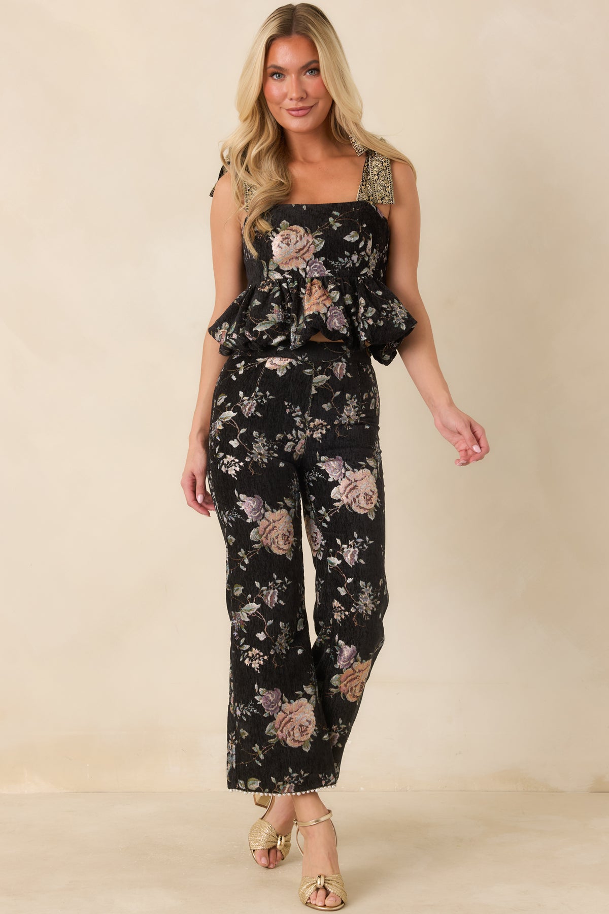 Black floral cropped pants with muted blush, peach, lavender, and green tapestry-style detailing and flared hems.