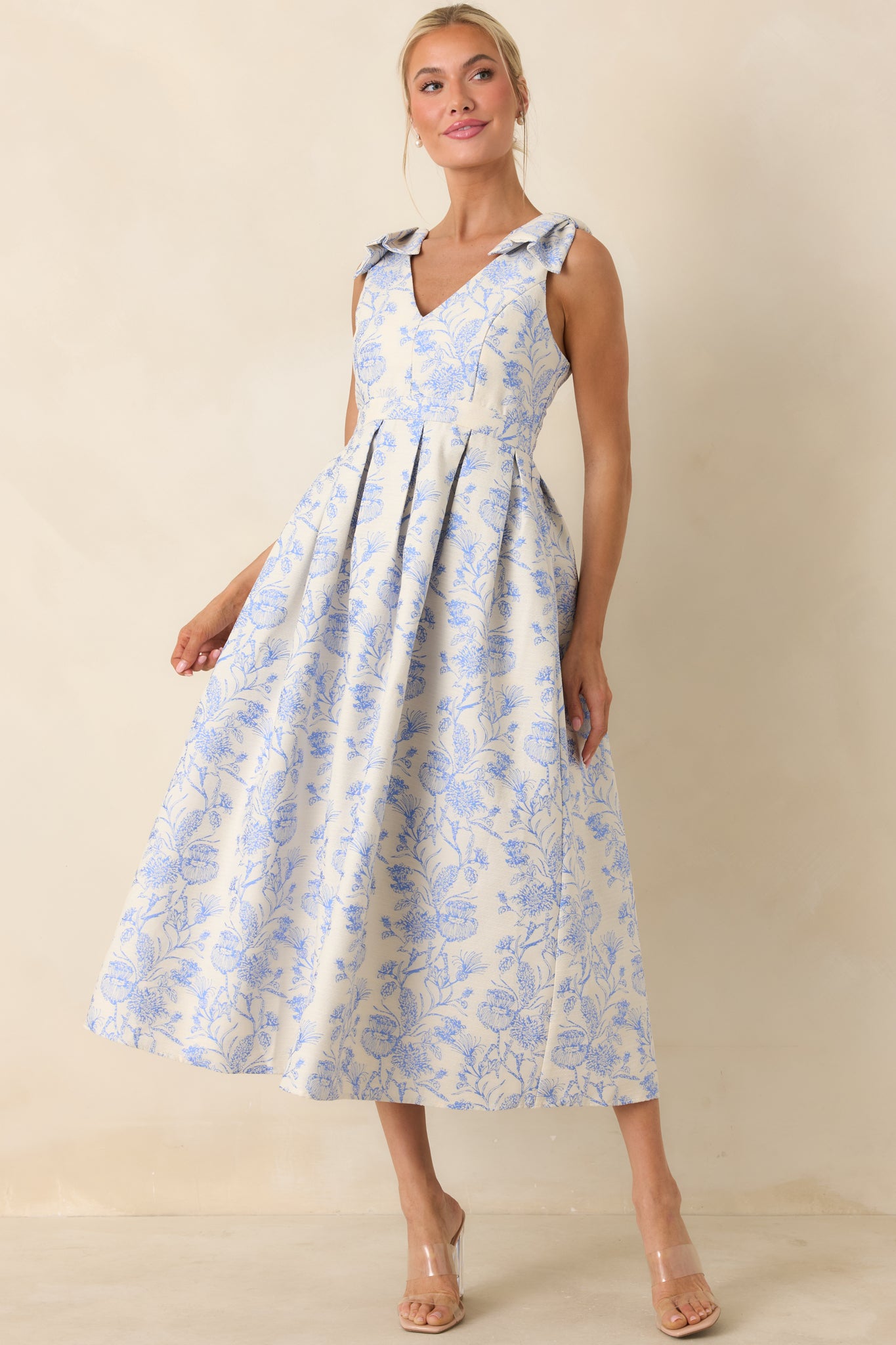 Blue floral midi dress with delicate bow straps and defined waist, shown from the front with a full pleated silhouette.