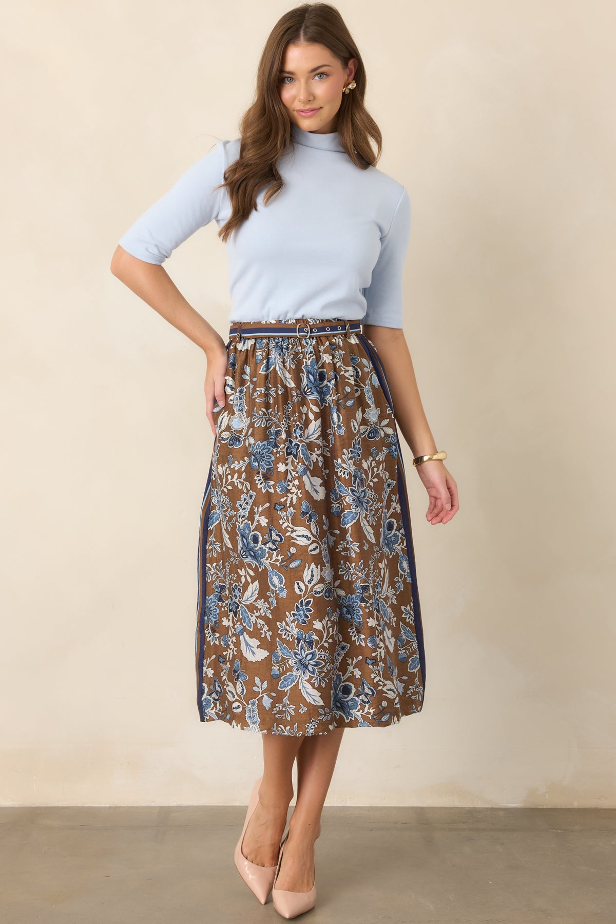 Brown A-line midi skirt adorned with blue floral motifs, a striped waistband and side border, double-buckle belt, and convenient side pockets.
