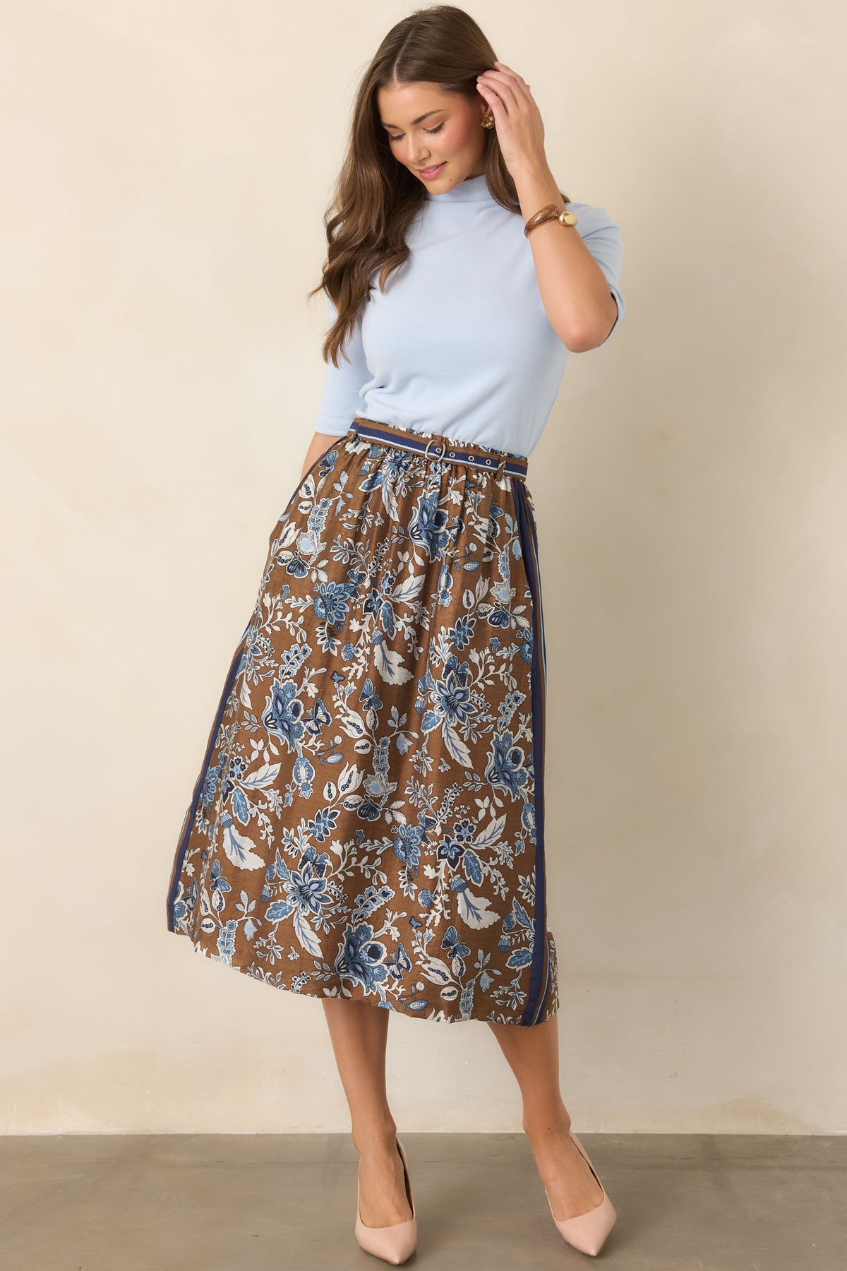 Floral-print brown midi skirt featuring layered blue blossoms, contrasting striped trim, a coordinating double-buckle waist belt, and softly structured A-line shape.