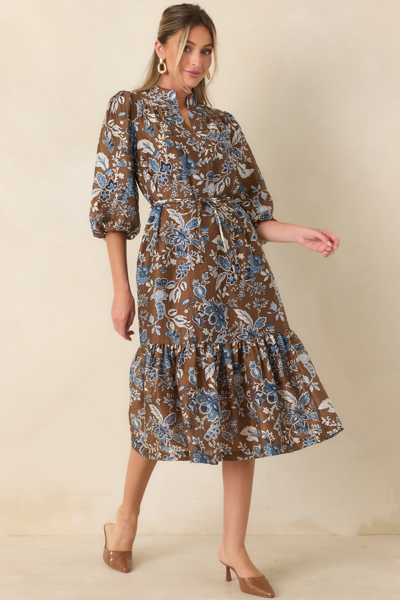 Brown floral midi dress featuring a blue botanical print, split band collar, three-quarter balloon sleeves, waist tie, and flowing ruffle skirt.