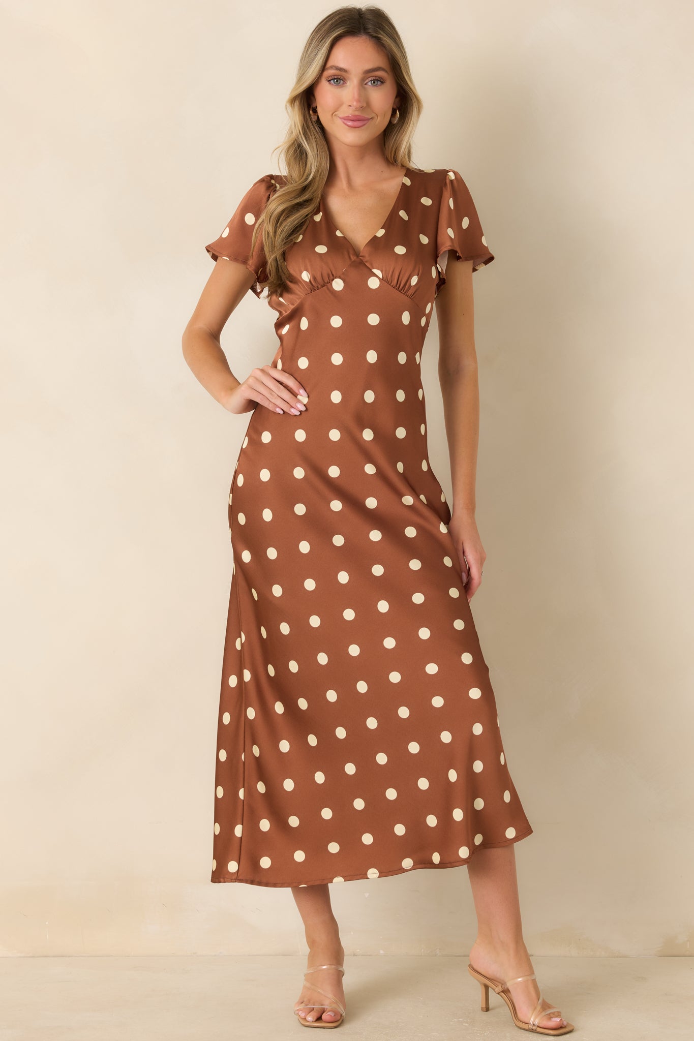 Brown satin midi dress featuring ivory polka dots, surplice neckline, flutter sleeves, and flowing flared skirt.