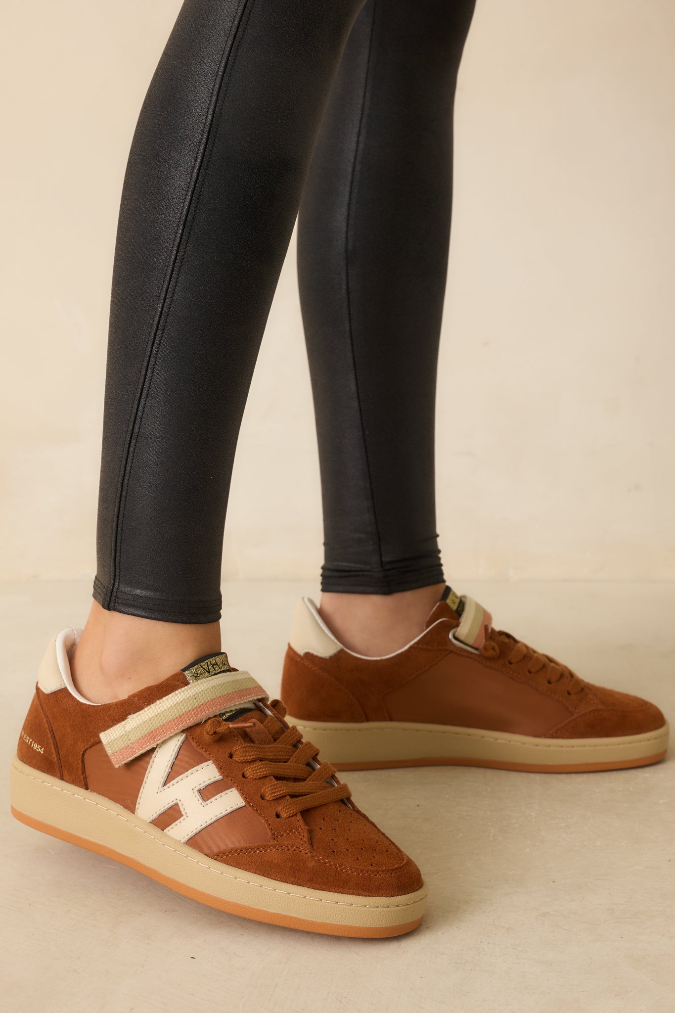 A side view of the brown low-top sneakers showing smooth leather and suede panels, striped velcro strap, and contrast side logo patch.