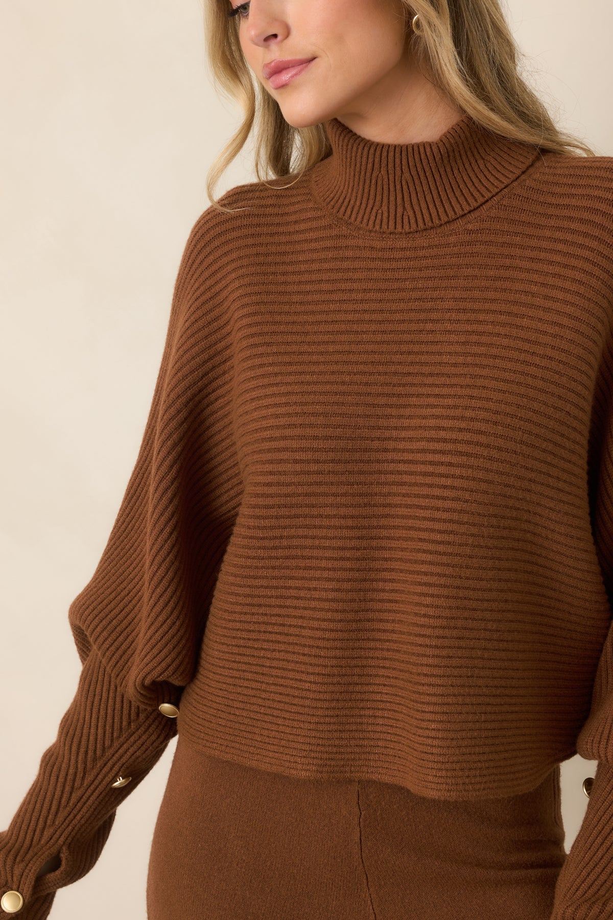 Close-up of the brown ribbed knit sweater highlighting the turtleneck collar, textured fabric, and button-accented cuff.