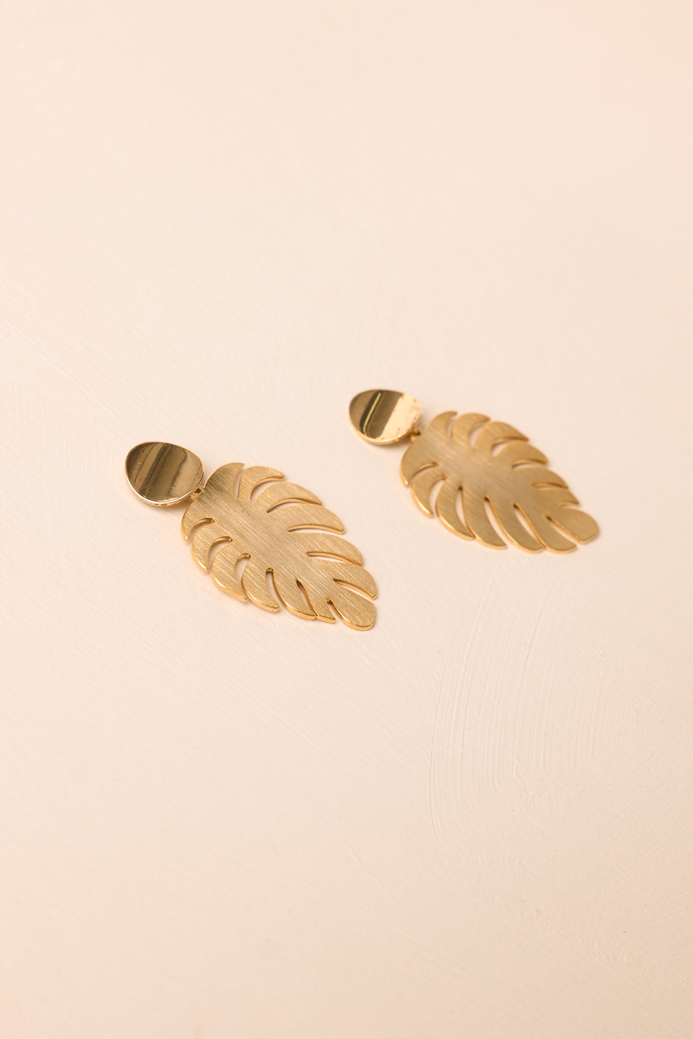 Brushed gold drop earrings with curved disc accents and a lightweight statement silhouette on beige.