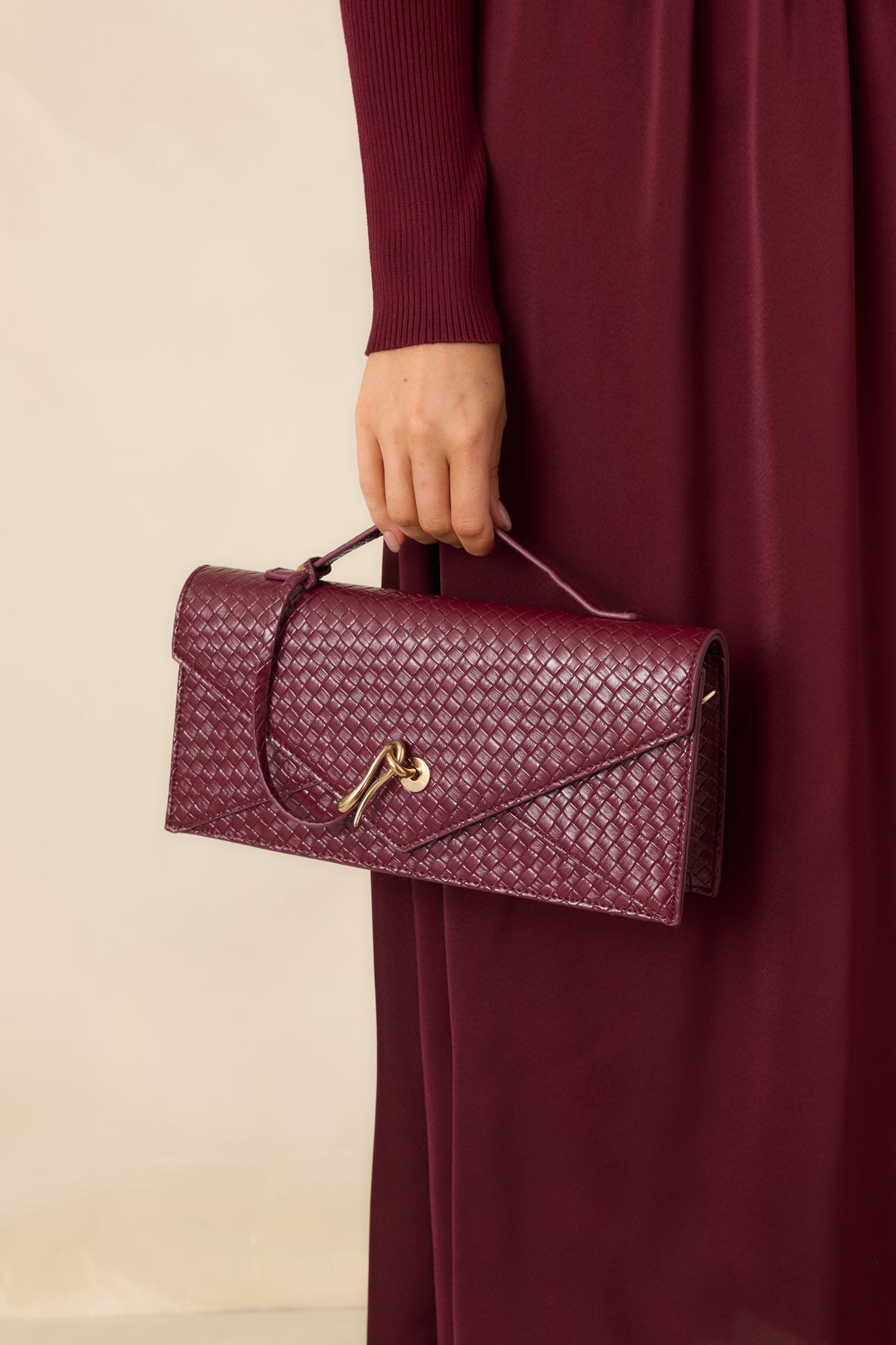 Burgundy faux leather clutch with a textured finish, pointed envelope flap, and sculptural gold-tone hardware.