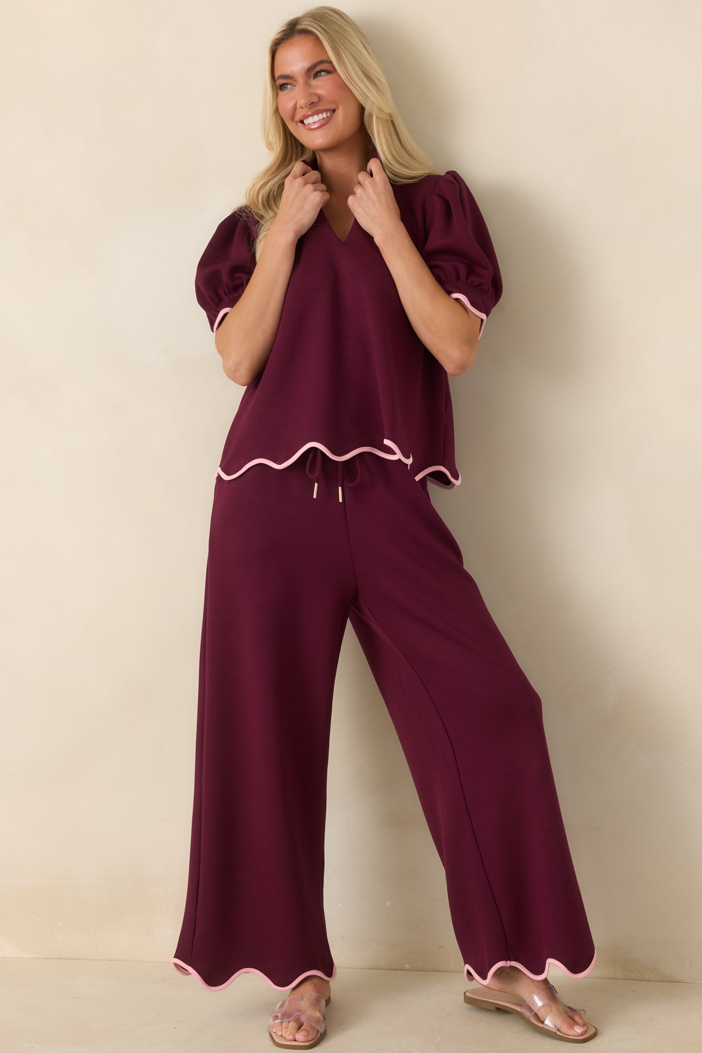 Burgundy knit top featuring a split neckline, modern stand collar, and blush pink scalloped trim along the sleeves and hem for a refined contrast.