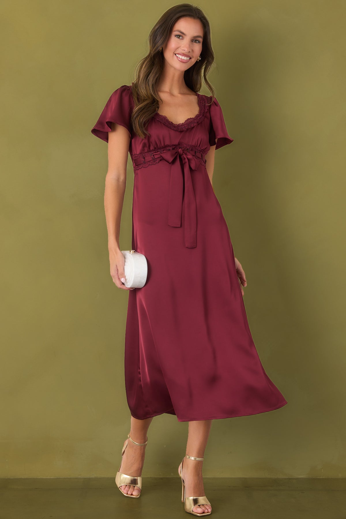 Rich burgundy satin midi dress featuring a scalloped lace neckline, flutter sleeves, and a gathered empire waist on olive green background