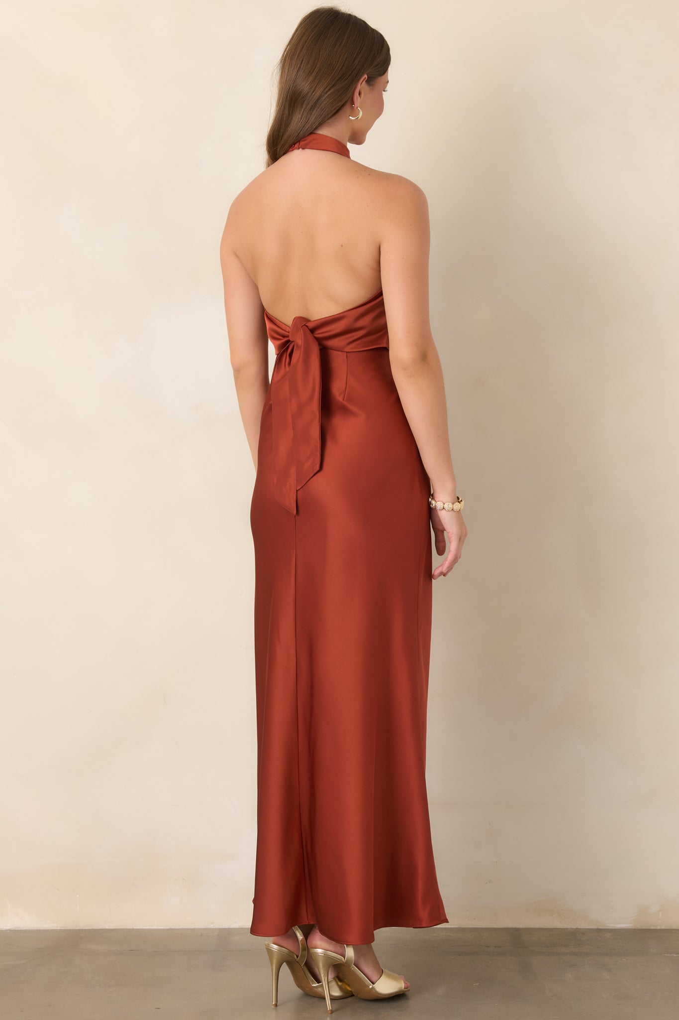 Rear view of the burnt sienna satin maxi dress showcasing its open back, dramatic self-tie detail, and fluid floor-length skirt.