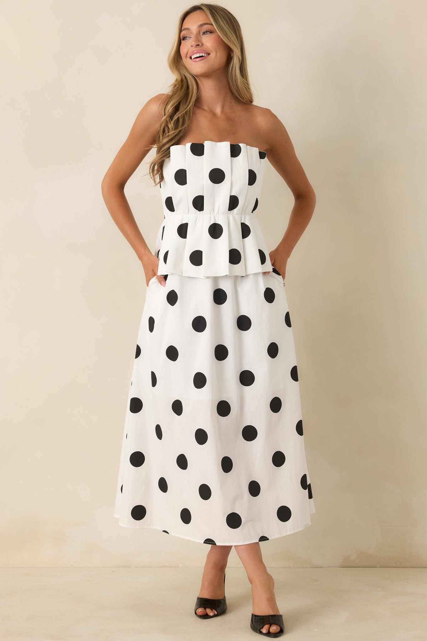 Chic black and white polka dot midi dress featuring a strapless neckline, tailored pleated waist detail, hidden side pockets, and an airy A-line shape.