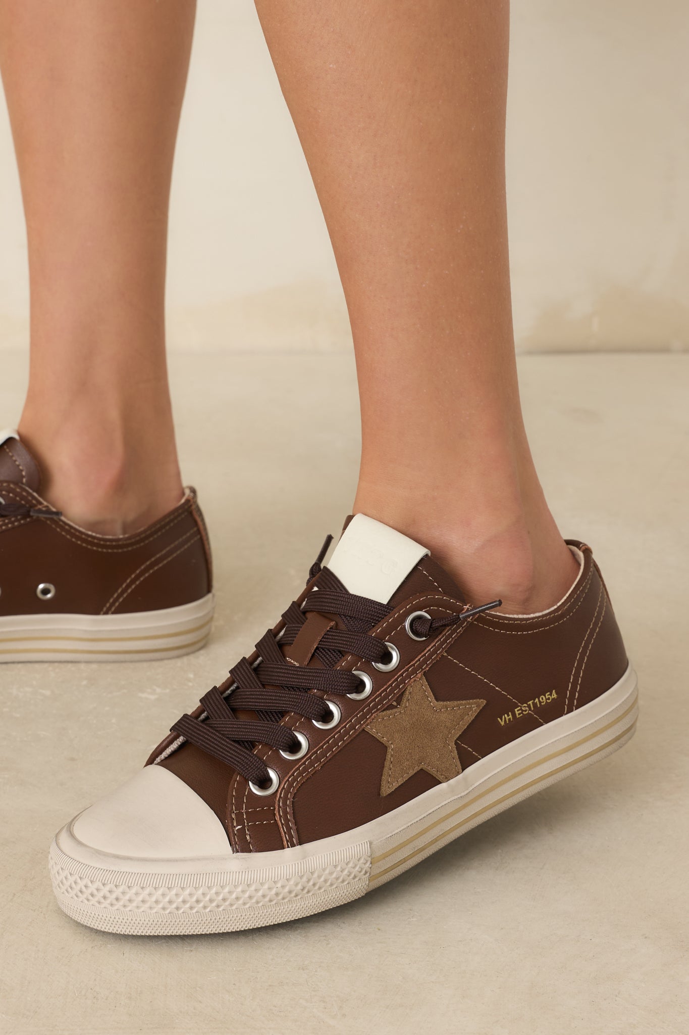 Chocolate brown low-top sneakers featuring a taupe suede star, ivory rubber toe cap, dark laces, and a layered vintage-inspired sole.