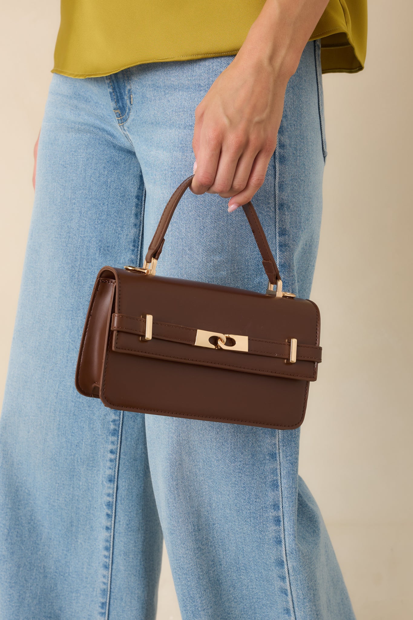Chocolate brown faux leather handbag featuring a structured silhouette, gold-tone hardware, top handle, and front belt detail.