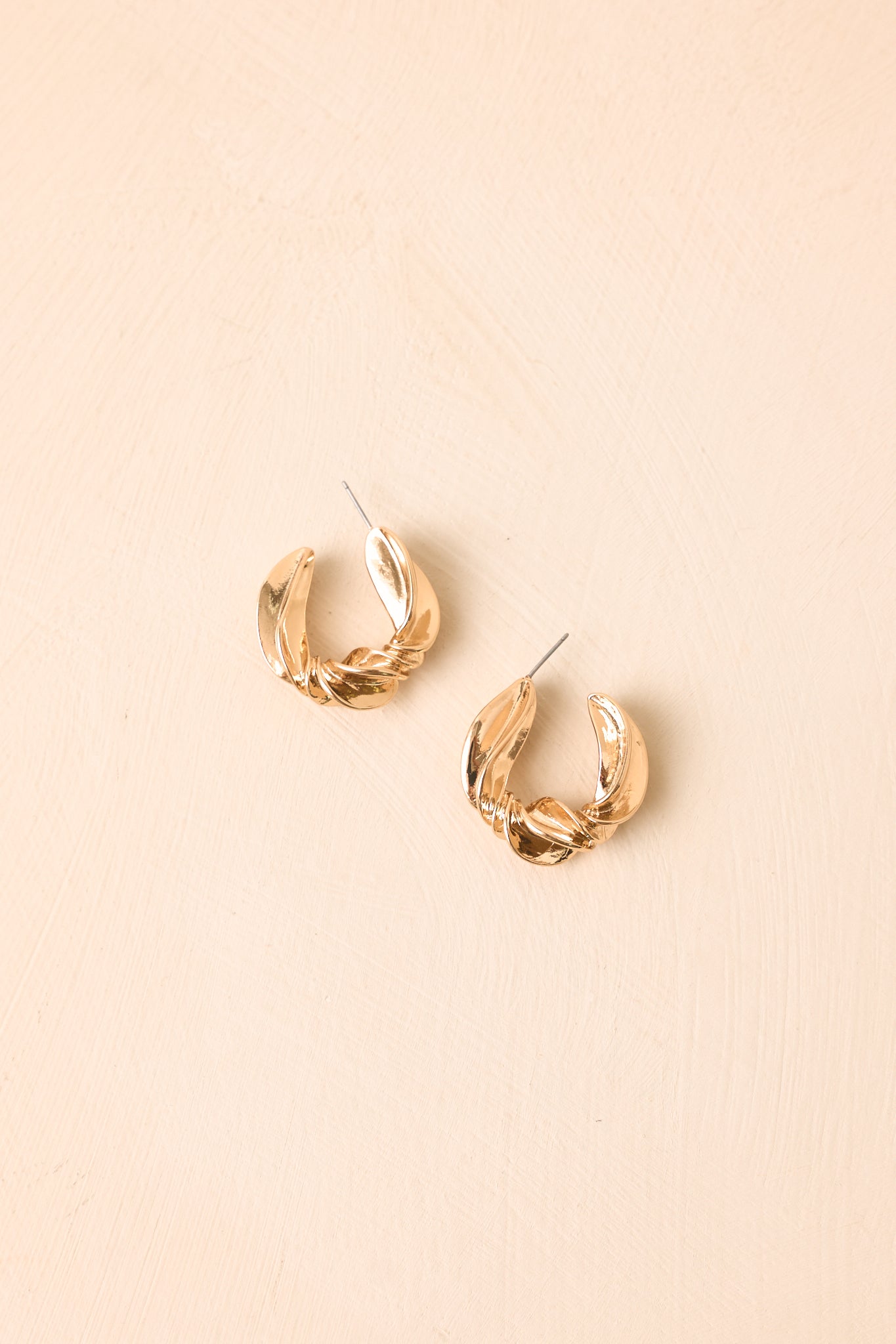Chunky gold-tone earrings with an artistic wrapped design presented on a neutral beige backdrop.
