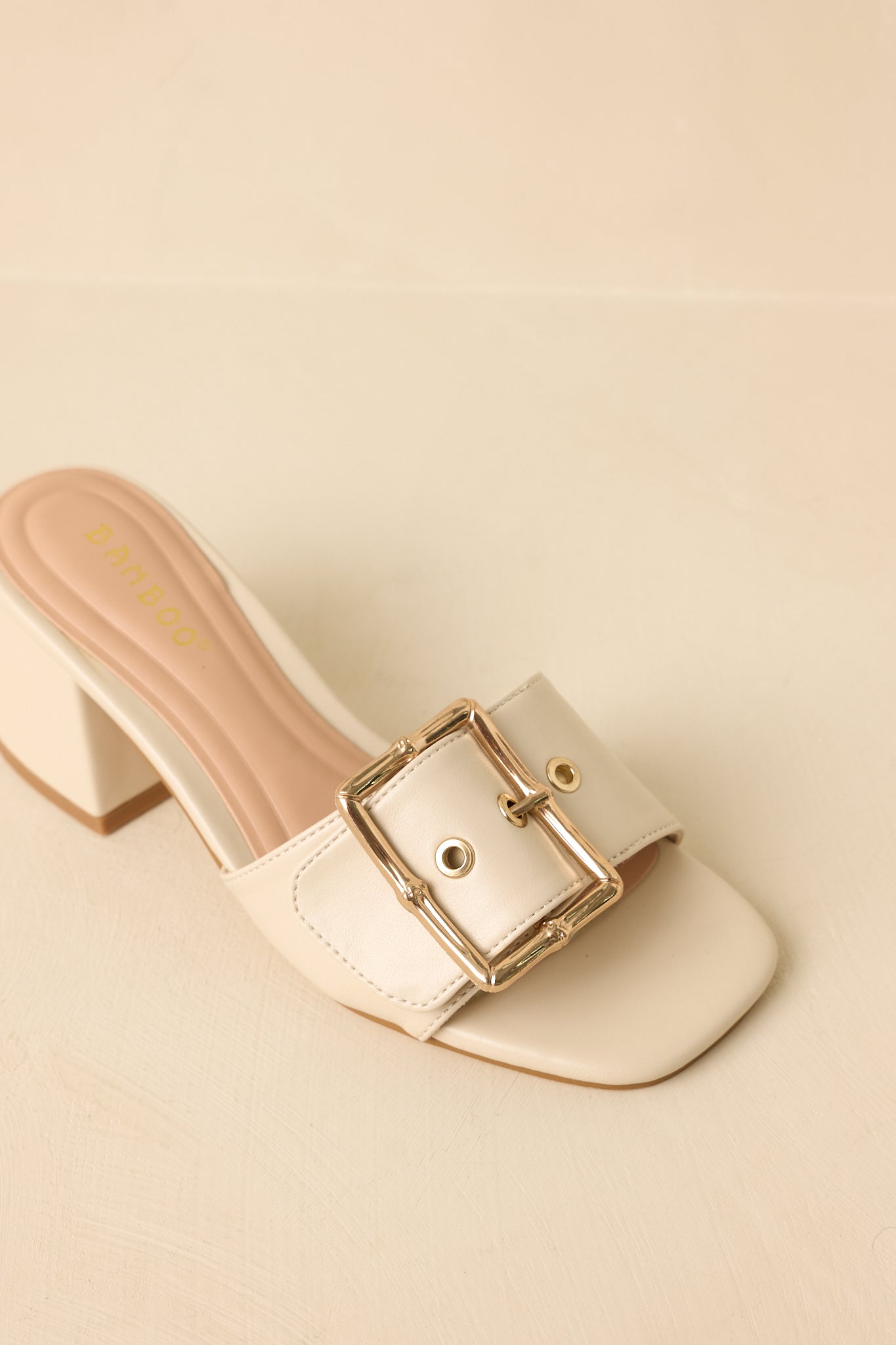 Close-up detail of the light beige block heels showing the gold-tone buckle hardware and smooth strap finish.