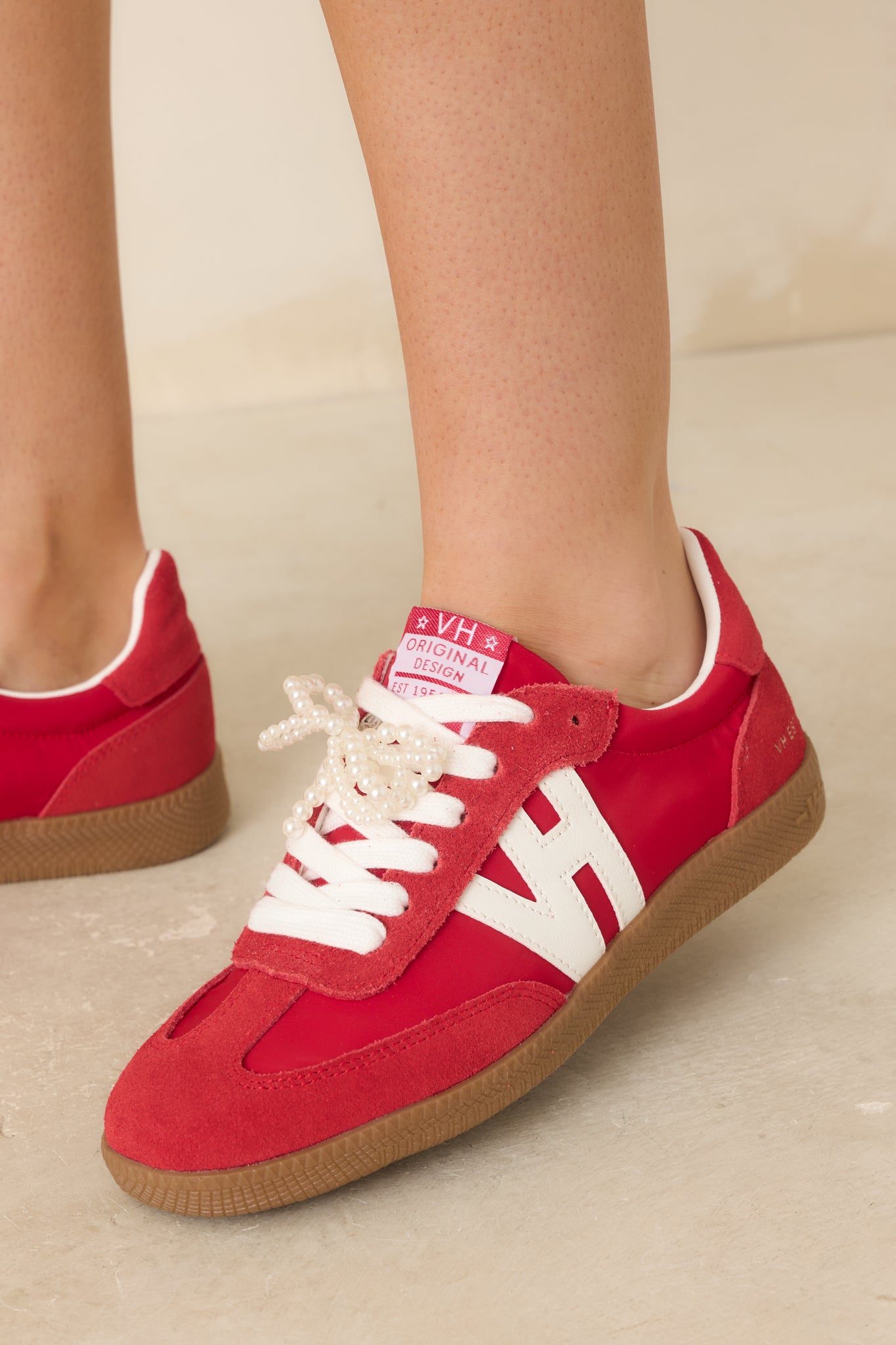 Close-up detail of red sneakers showing removable pearl bow, white laces, and textured mixed materials.