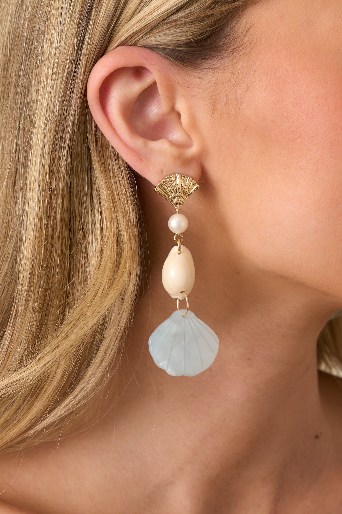 Close-up photo of earrings that features a drop design with a gold seashell stud, ivory faux pearl charm, ivory seashell charm, and a light blue pearlescent seashell bottom charm.