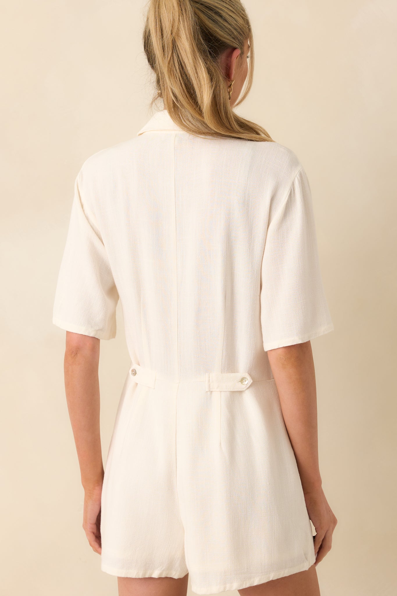 Up-close photo of back of model wearing ivory romper that features a lightweight rayon-linen blend fabric, collared neckline, functional button front, and short sleeves.