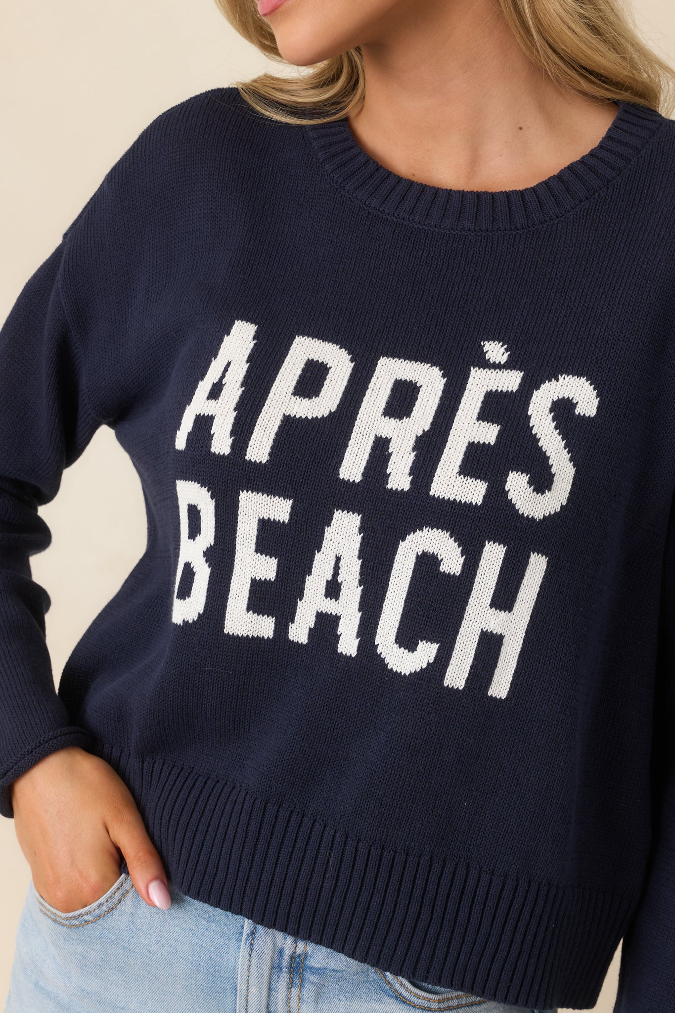Close-up of the navy sweater showing ribbed trim, smooth knit texture, and “Après Beach” lettering knit directly into the fabric.