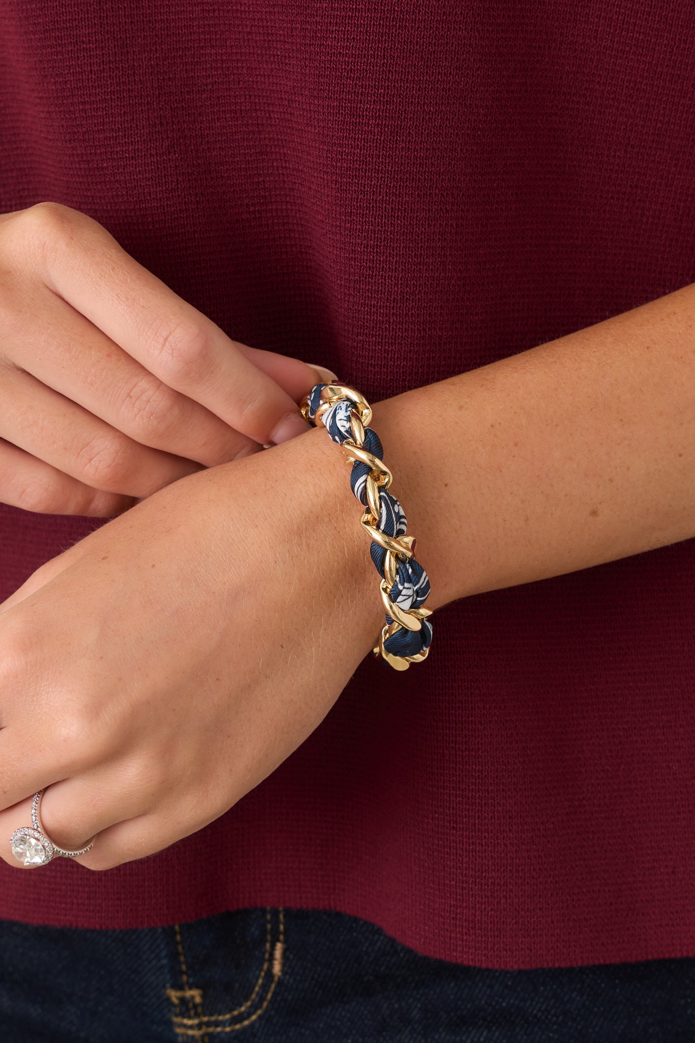 A close-up of a person's wrist wearing a gold link and blue scarf braided bracelet, paired with a burgundy top and dark blue jeans.