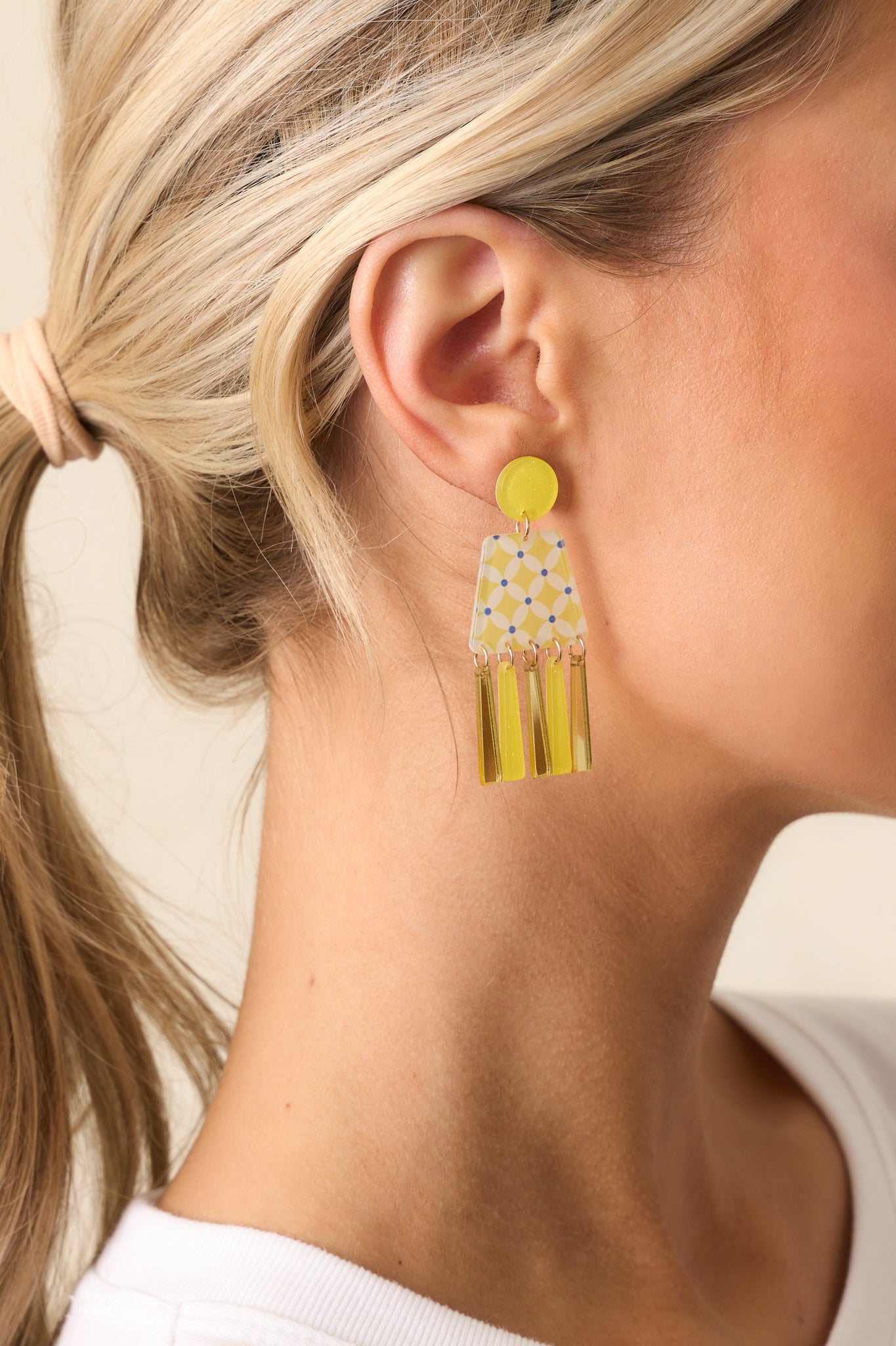 Close-up of a geometric yellow acrylic earring showing the glitter circle stud and connected clear trapezoid with yellow and blue print.