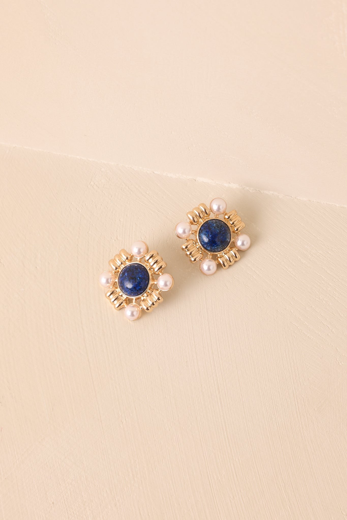 A close-up of earrings capturing the marble-like texture of the navy stone, the luster of the faux pearls, and the intricate gold accents on a beige background.