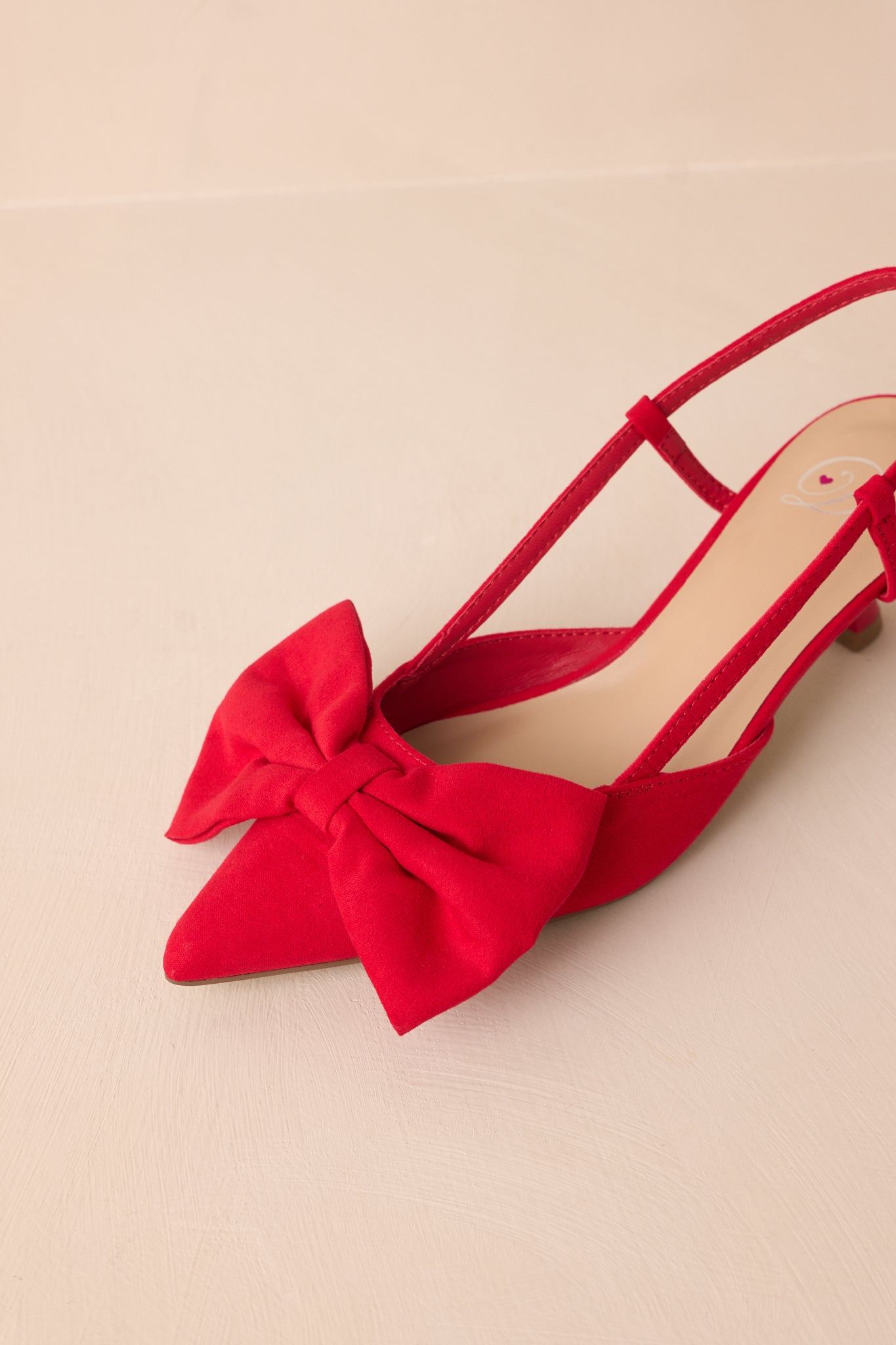 Close-up of the red kitten heels' pointed toe, emphasizing the texture of the material and the oversized red bow in sharp focus.