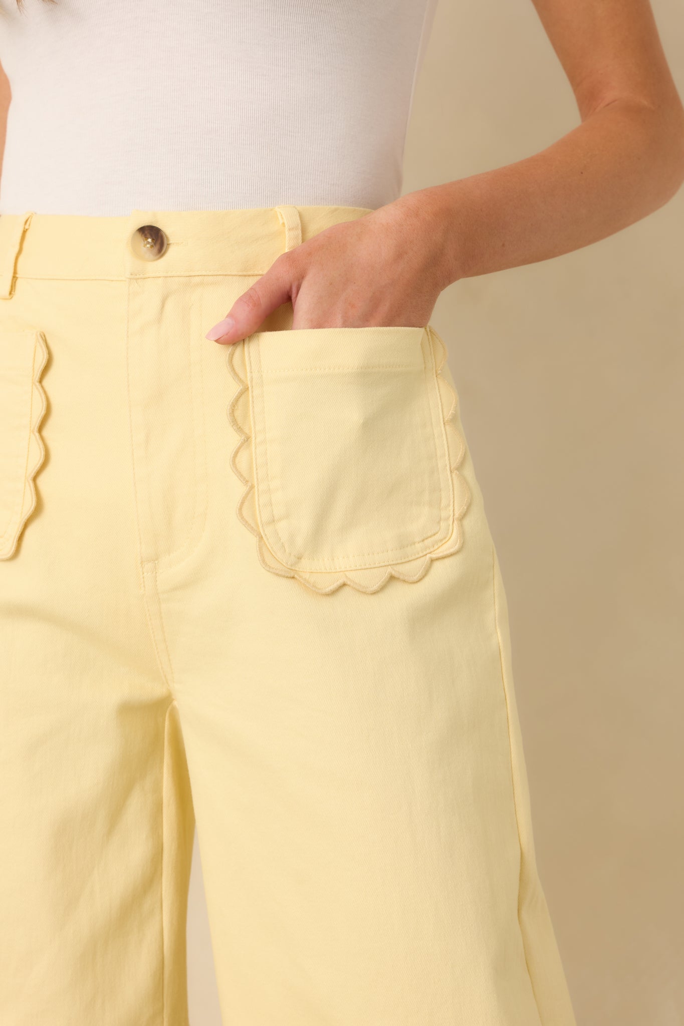 Close-up of yellow cotton pants showing scalloped front patch pockets, button closure, and waistband.