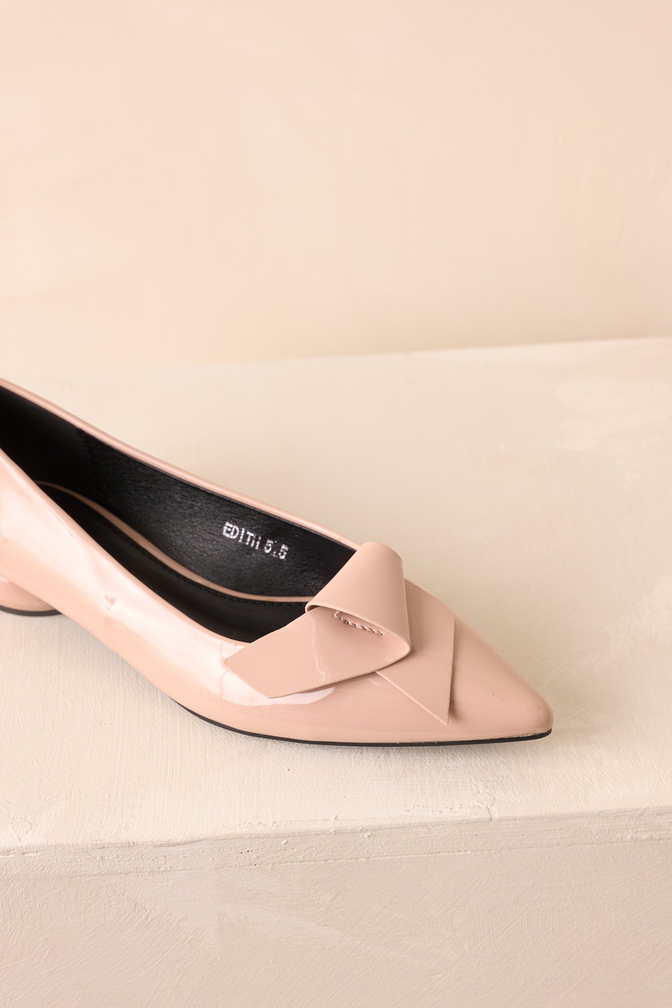Close-up of the beige flats, showing the bow detail, low sculpted geometric heel, and glossy material.