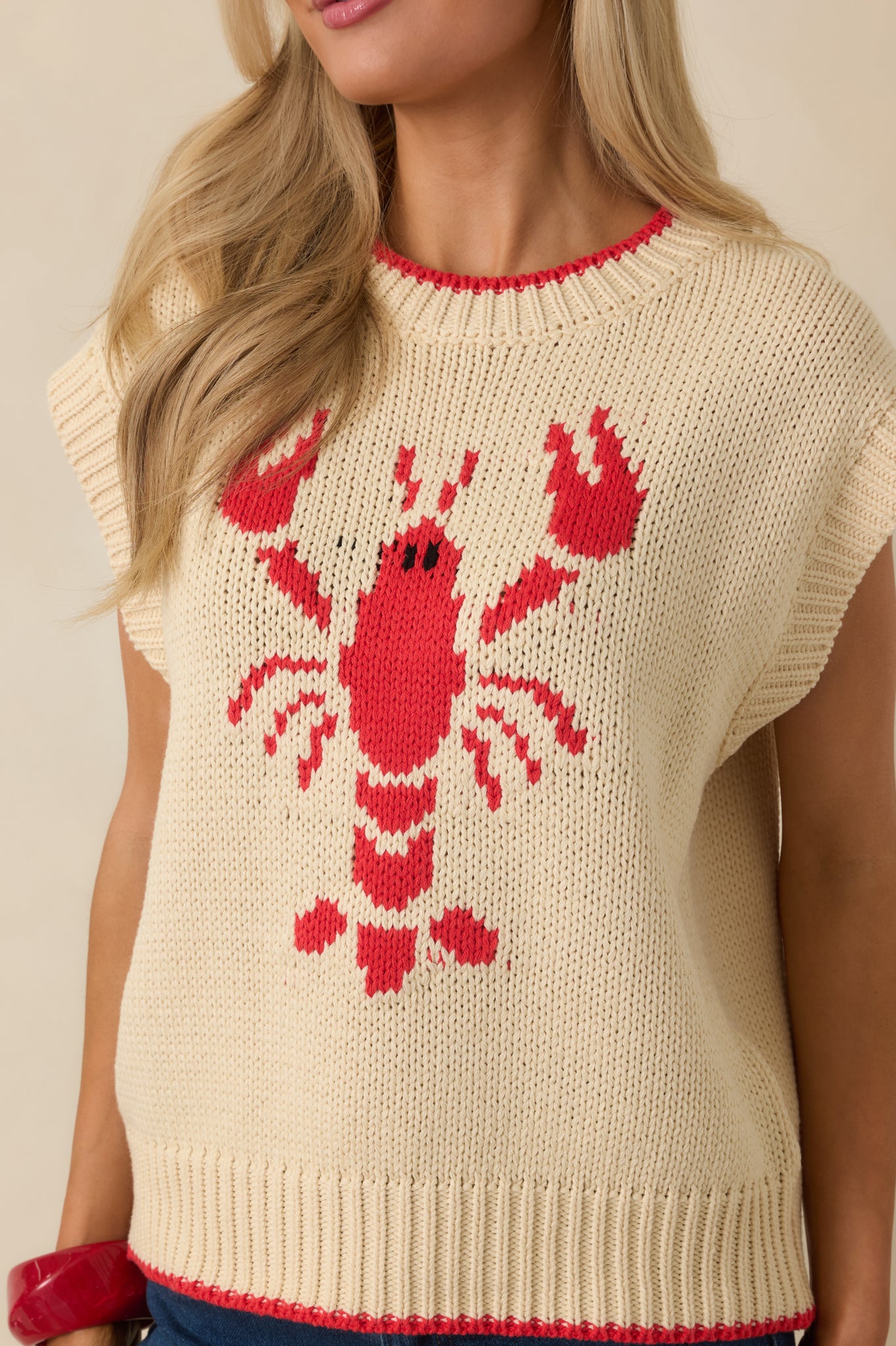 Close-up of the ivory sleeveless sweater showing the embroidered lobster motif and ribbed neckline detail.