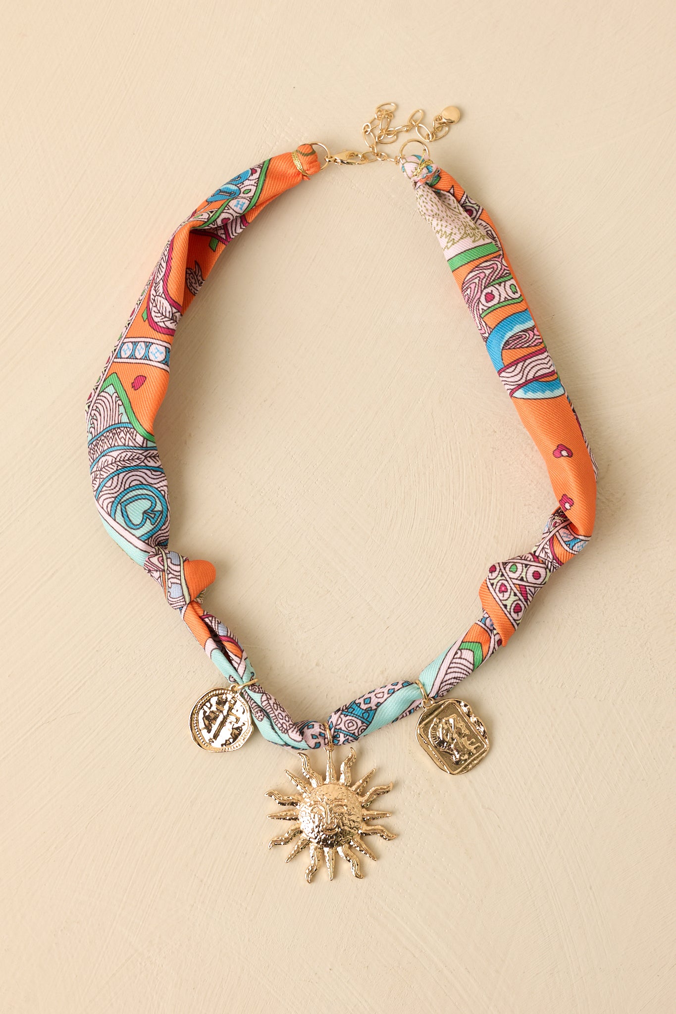 Colorful scarf necklace displayed against a beige background featuring orange tones and mixed gold charm accents.