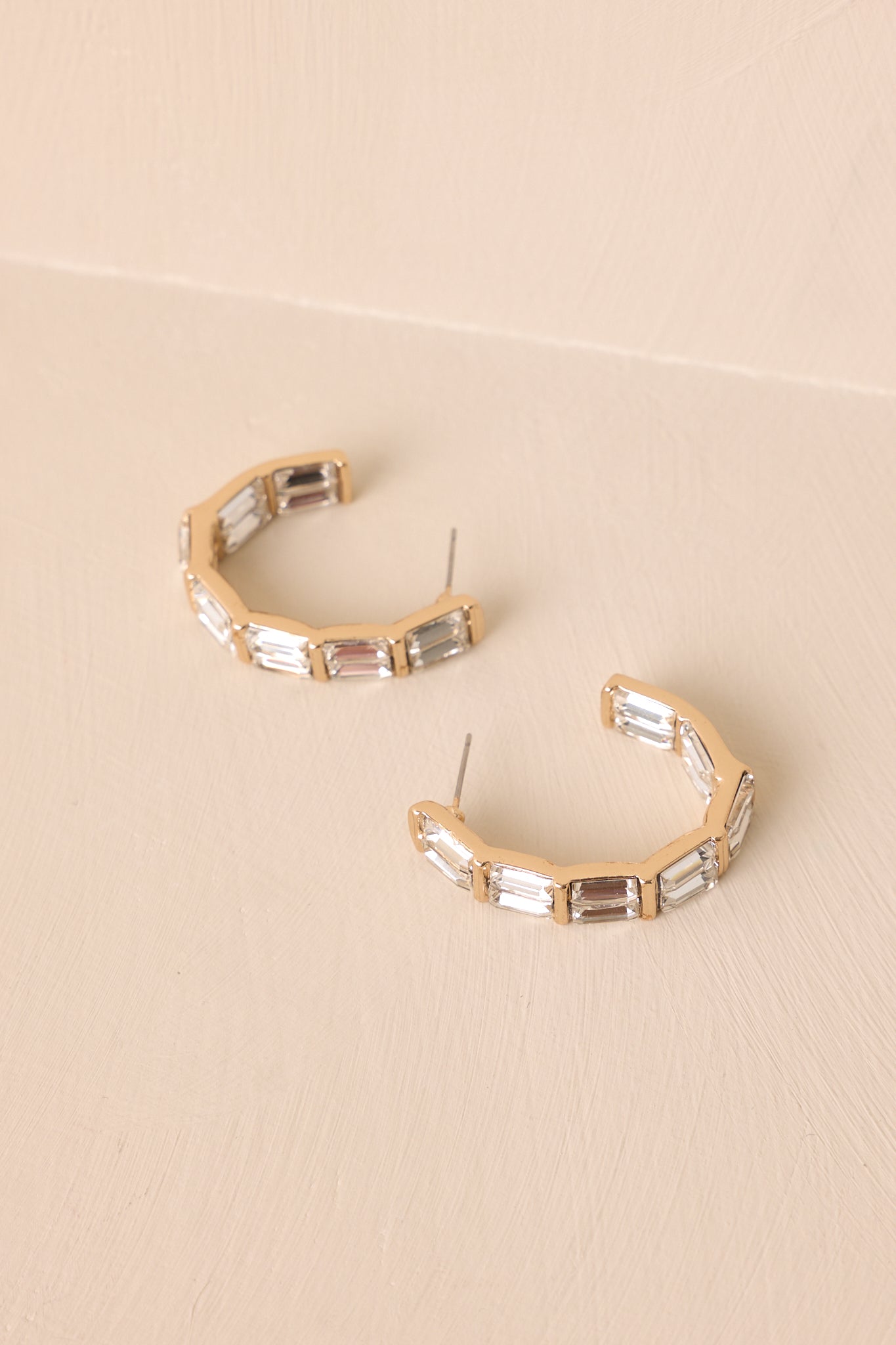 The gold and rhinestone earrings displayed against a neutral beige background, showcasing their curved shape and elegant design
