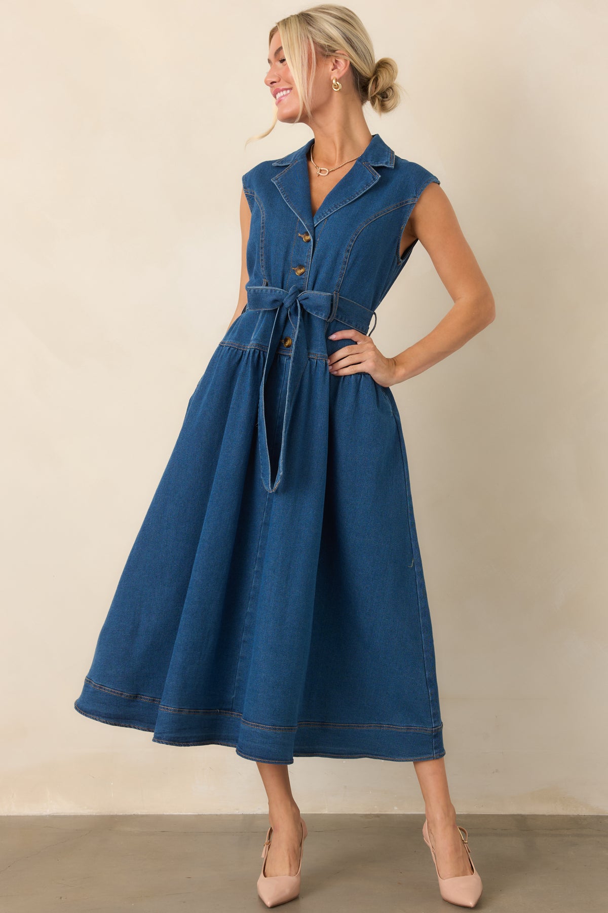Dark wash denim midi dress showcasing its sleeveless design, pointed collar, gold-tone button-front closure, cinching self-tie belt, and convenient side pockets.
