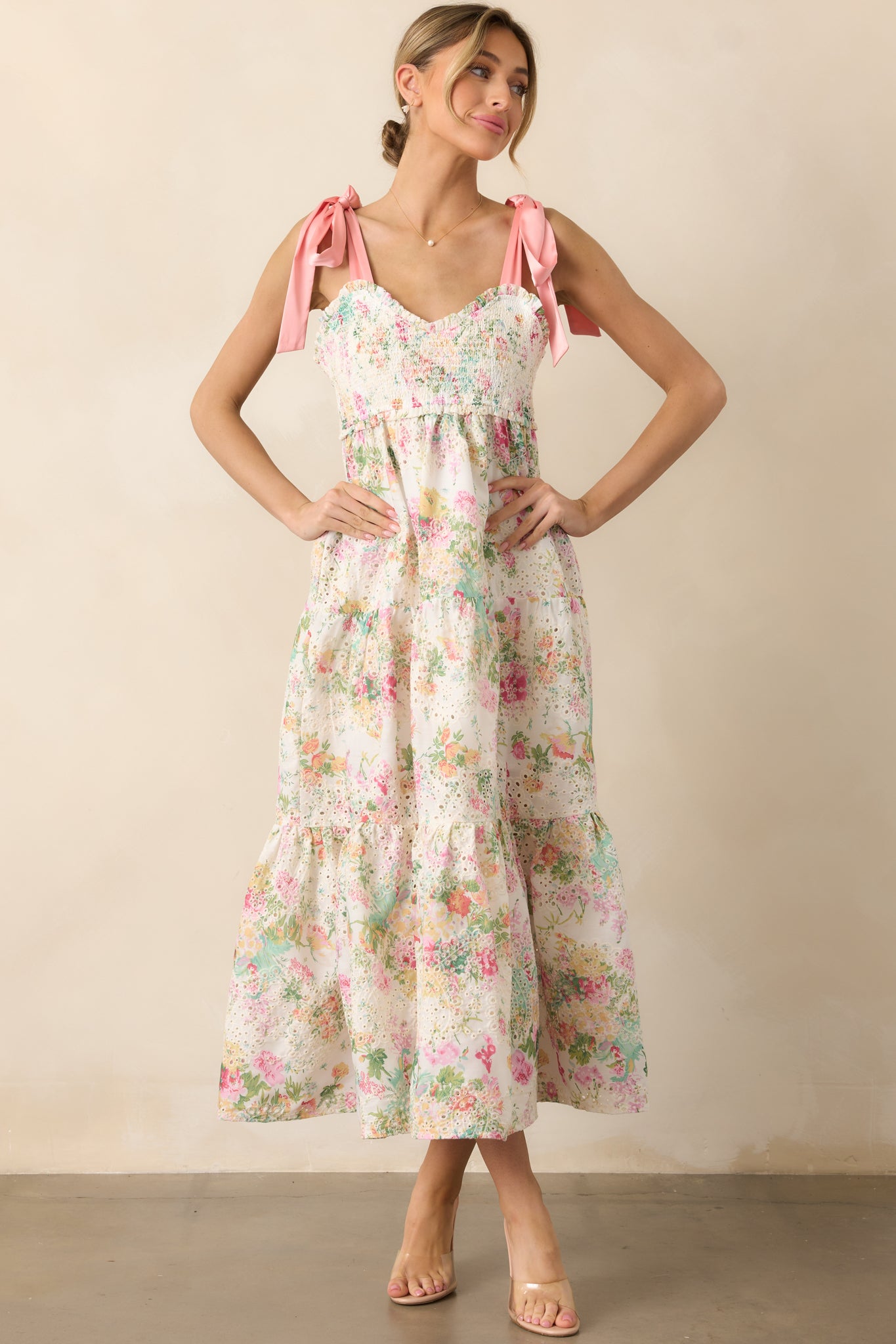 A beautiful light pink dress with multicolor floral print, a sweetheart neckline, self-tie satin bow straps, and a fully smocked bust, enhanced by embroidered eyelet detailing and a tiered skirt with functional pockets.