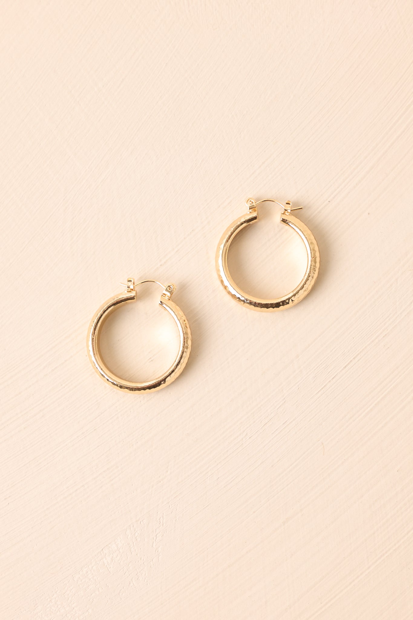 Flat lay of the earrings on a beige surface showing the reflective hammered surface and secure latch closure.