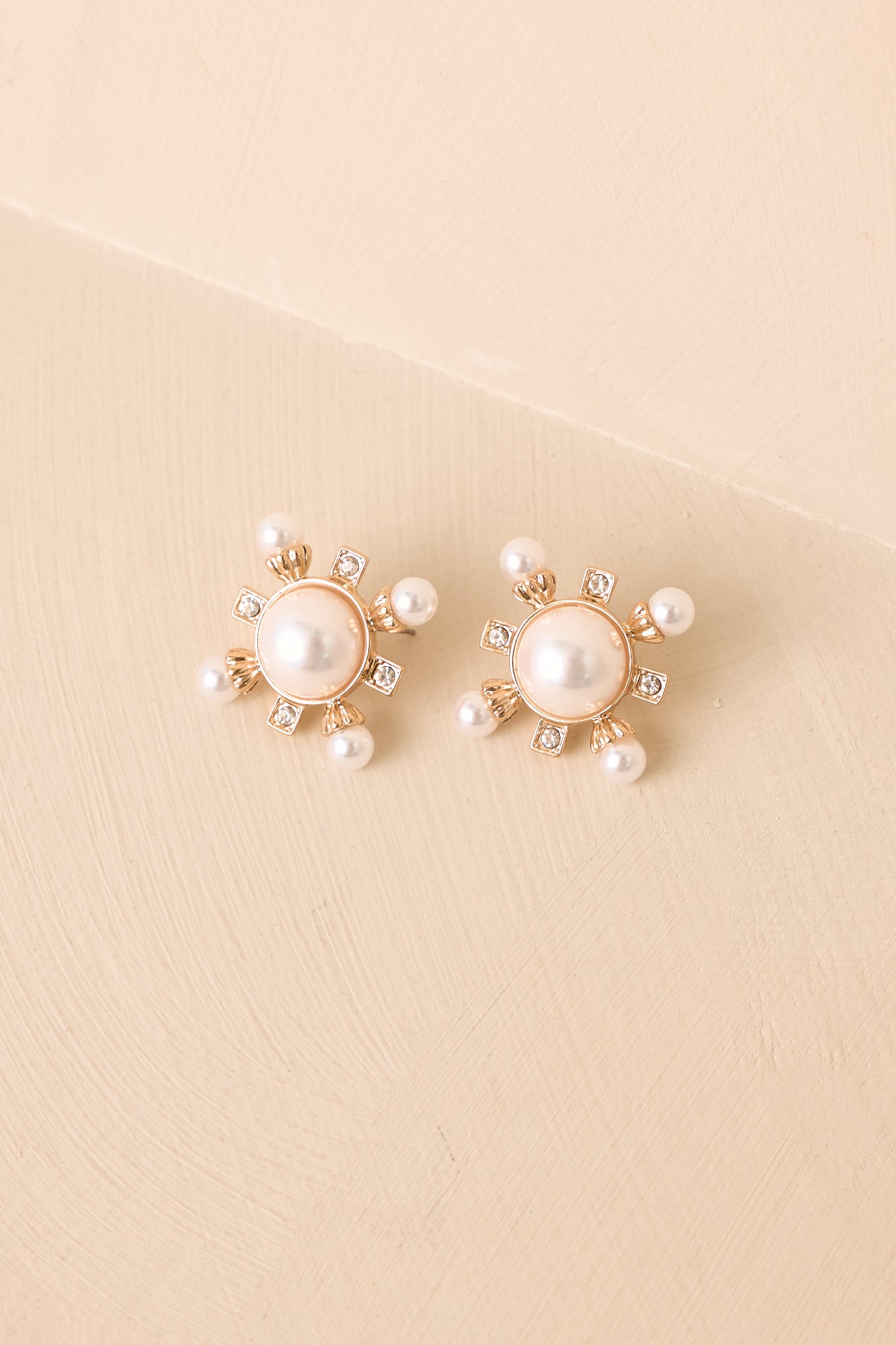 Faux pearl and crystal stud earrings displayed against a beige background with gold-tone details.