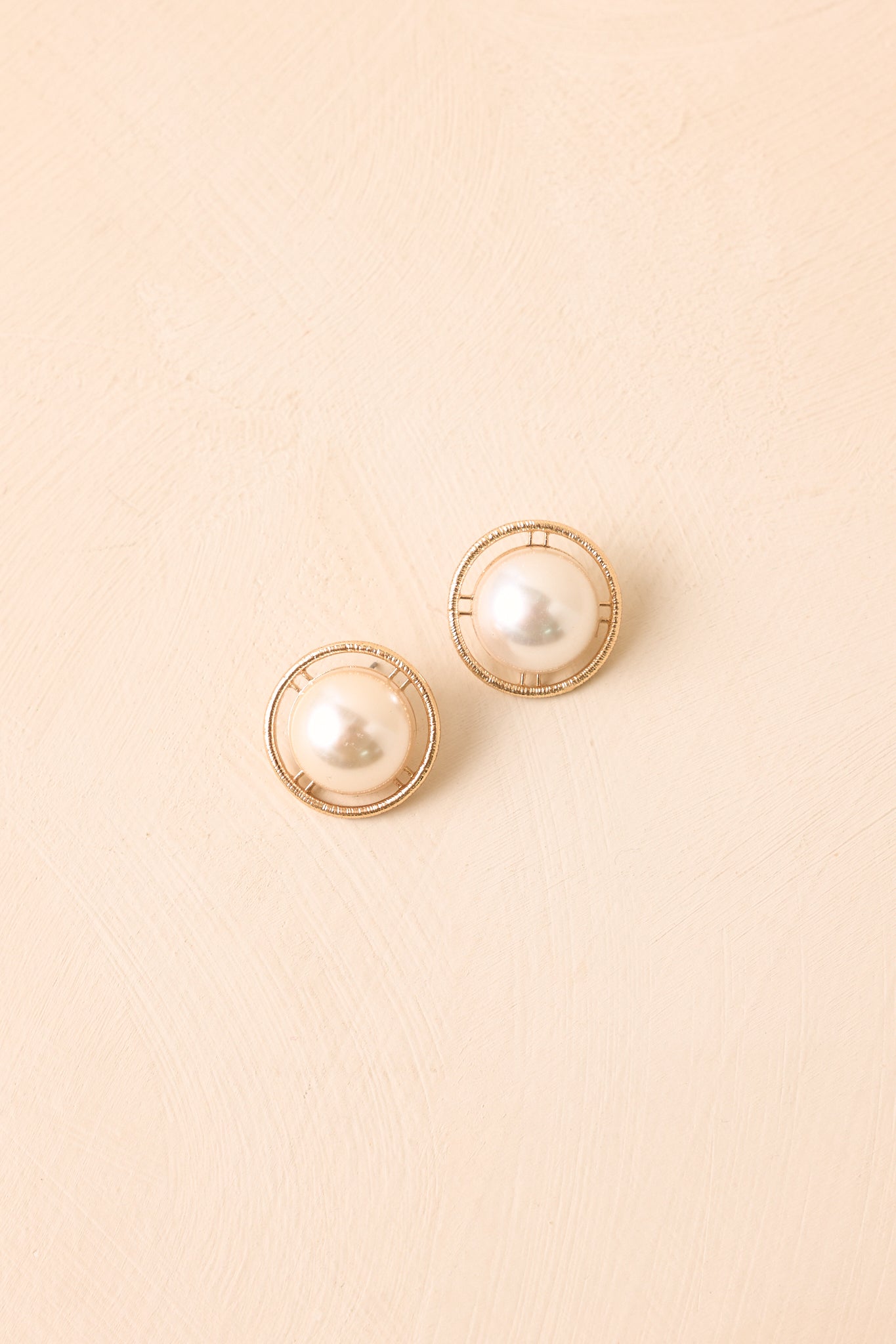 Faux pearl button earrings displayed against a beige background, highlighting the textured gold-tone halo and classic shape.