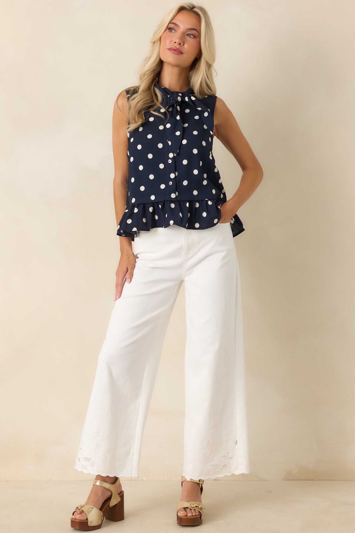 Feminine polka dot blouse featuring a button-front design, delicate neckline tie, sleeveless cut, and flirty ruffle finish.