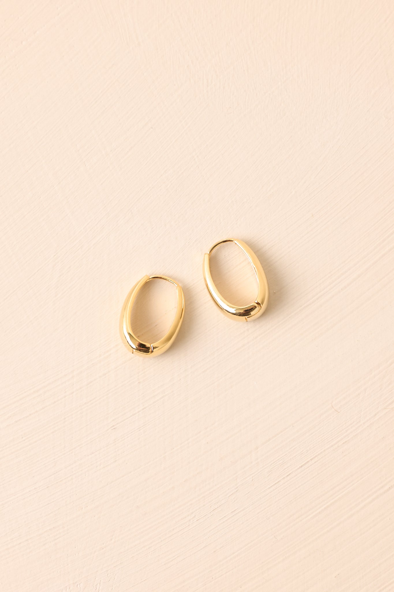Flat lay of the earrings on a beige surface showcasing the clean oval silhouette and polished gold tone.