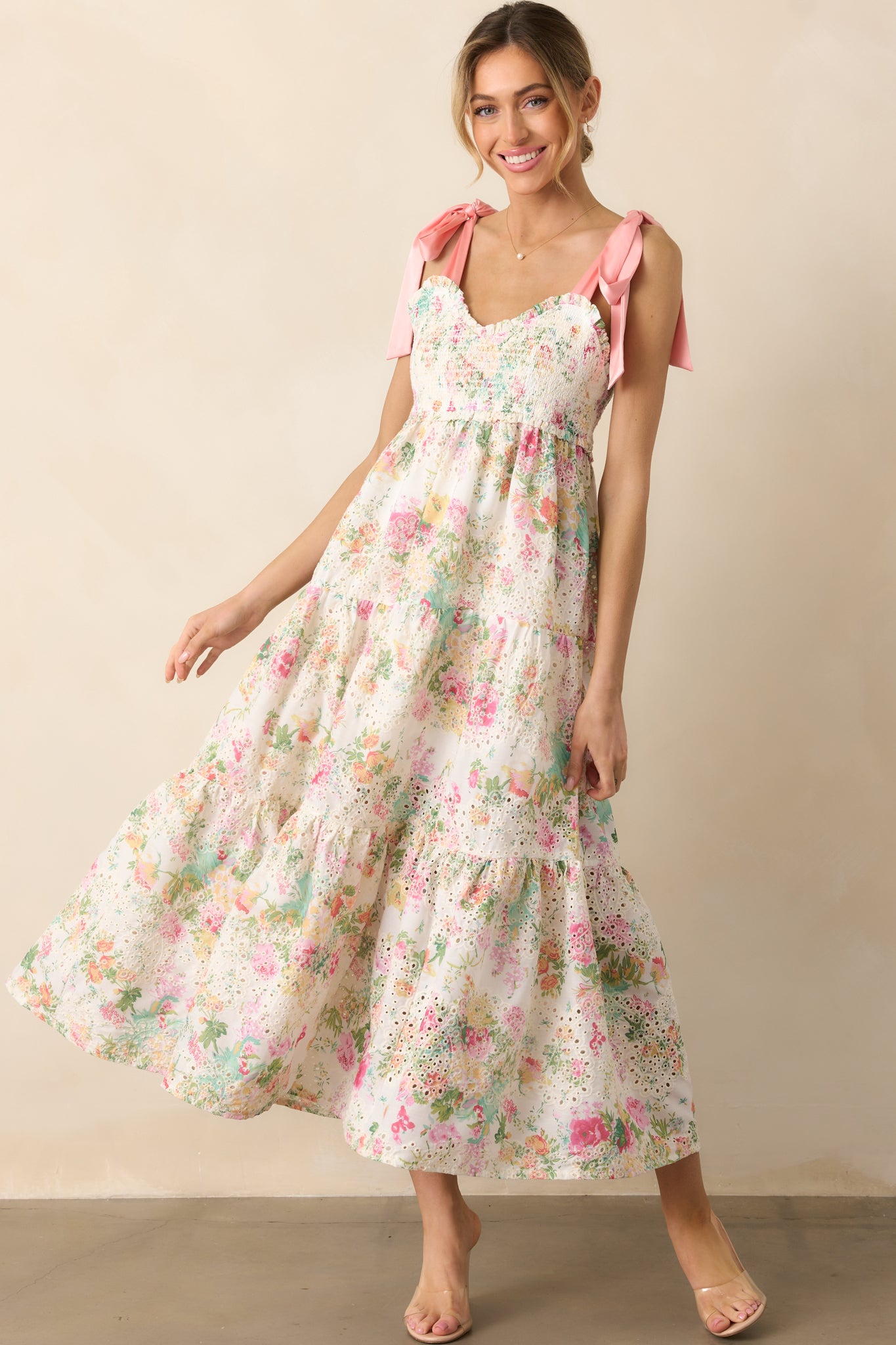A feminine light pink dress with a multicolor floral print, a sweetheart neckline, embroidered eyelet detailing, and a tiered skirt, finished with self-tie satin bow straps and functional pockets.