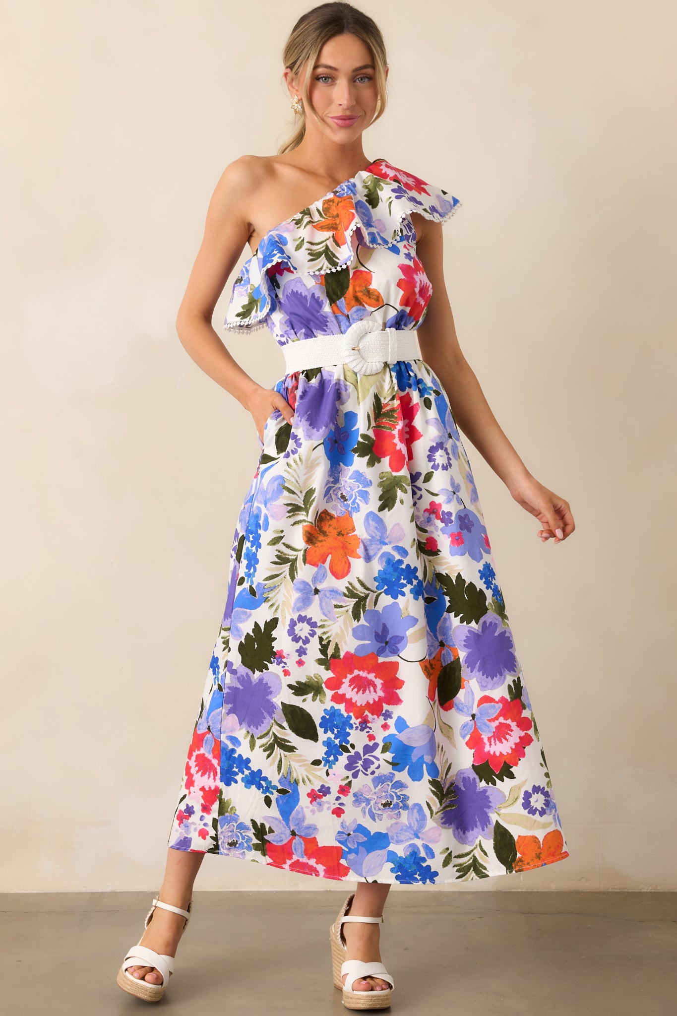 The floral maxi dress in white with blue and red blooms, featuring an elastic one-shoulder ruffle neckline, eyelet trim detailing, and an elastic waist, creating a flattering, feminine silhouette with functional pockets for added convenience.