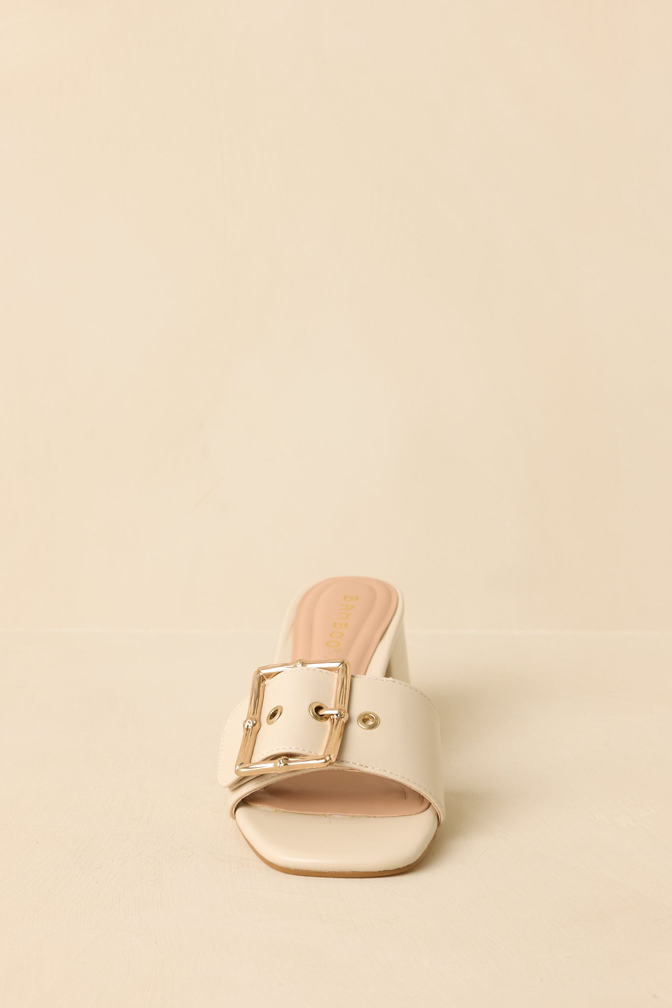 Front-facing view of light beige open-toe block heels against a beige background, highlighting the wide strap and buckle detail.