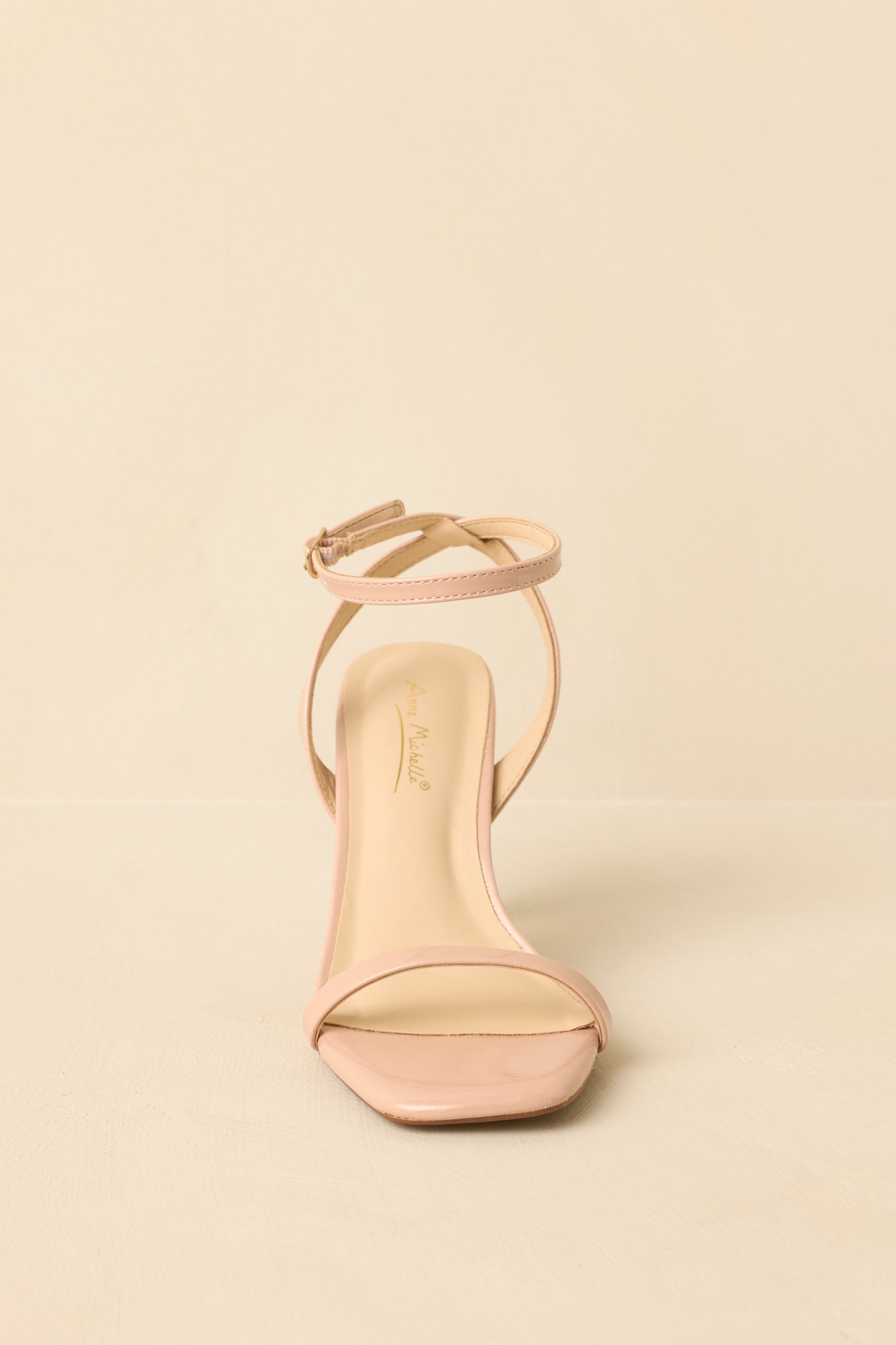 Front-facing view of nude open-toe heels against a beige background highlighting the slim toe strap.