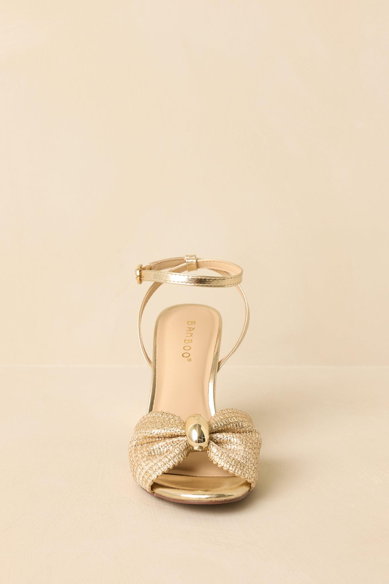 Front view of gold woven sandals showing the bow-shaped strap, open toe, and metallic block heel.