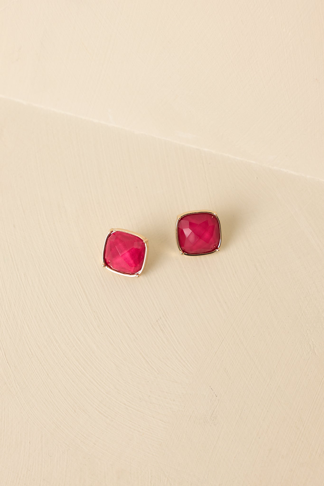 Front view of cranberry red stud earrings displayed on beige background emphasizing gemstone sparkle and gold frame.